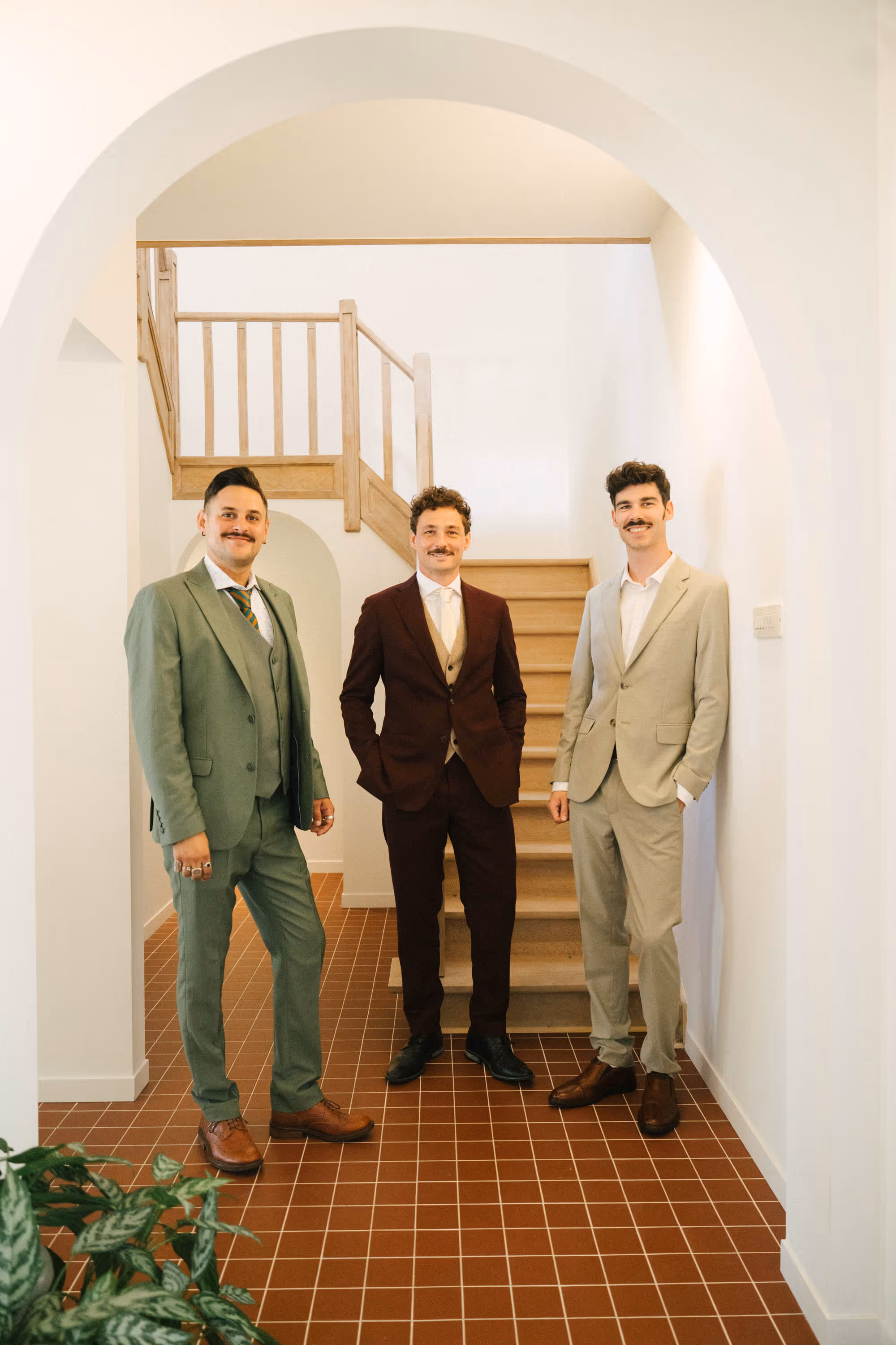 Three men with mustaches wearing suits standing indoors in front of a wooden staircase.
