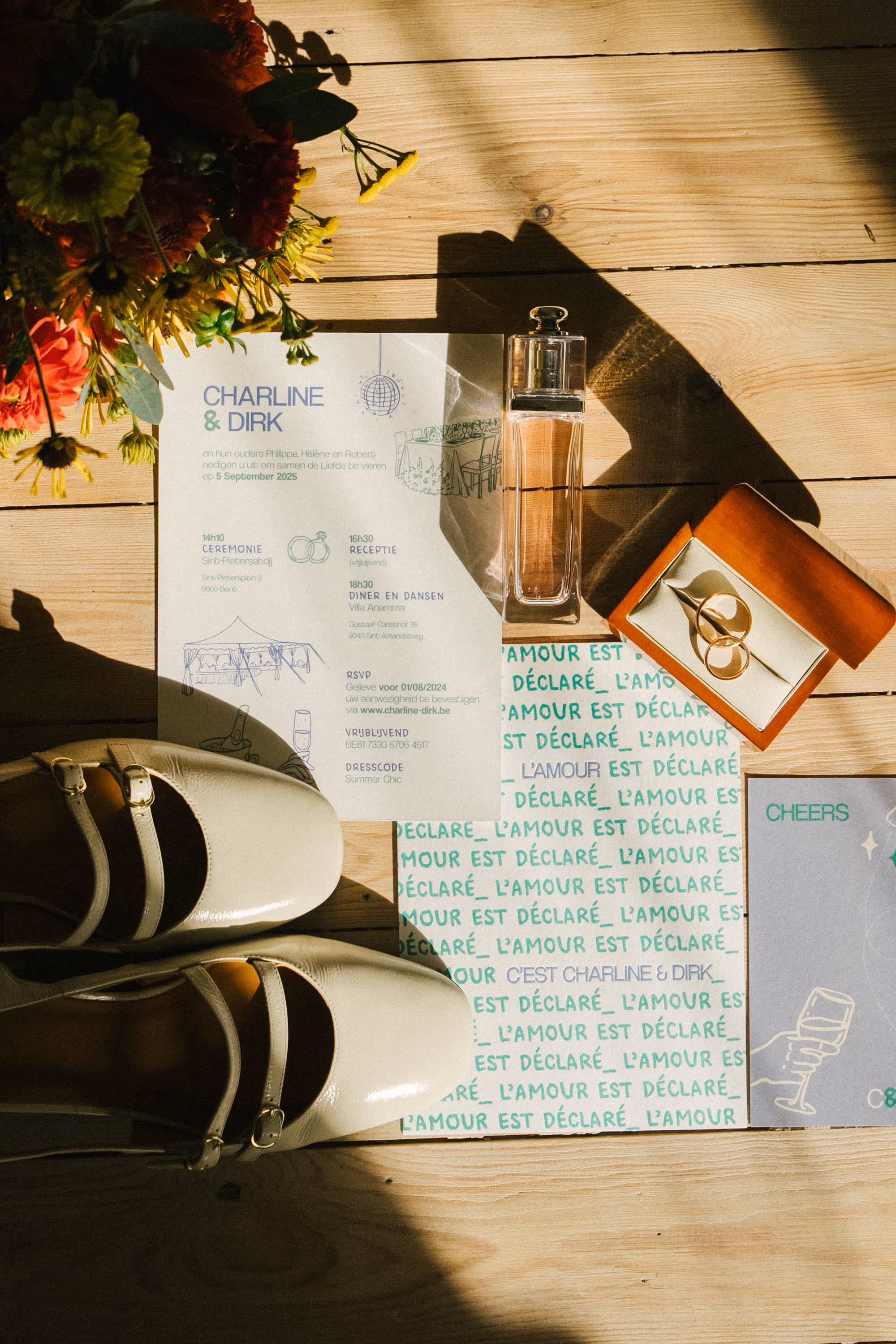 Wedding invitation flat lay with cream shoes, perfume bottle, wedding rings in wooden box, bouquet, and invitation cards on wooden surface.