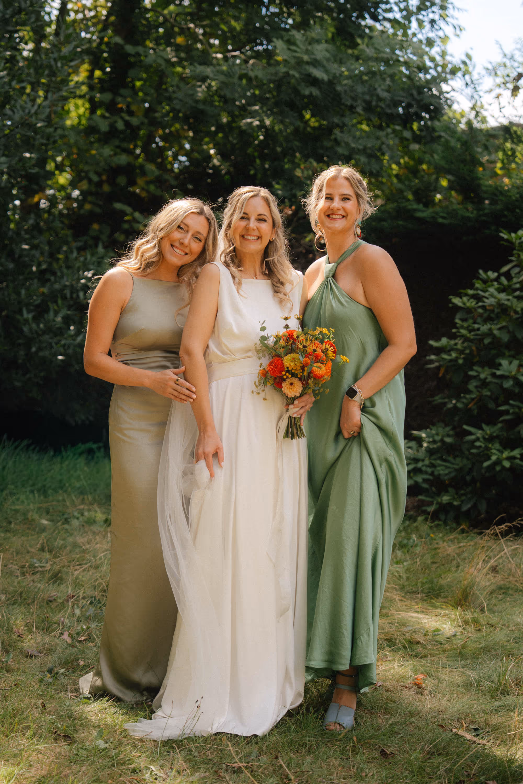 Three smiling women in formal dresses stand outdoors on grass with greenery in the background; the center woman in a white dress holds a colorful floral bouquet.