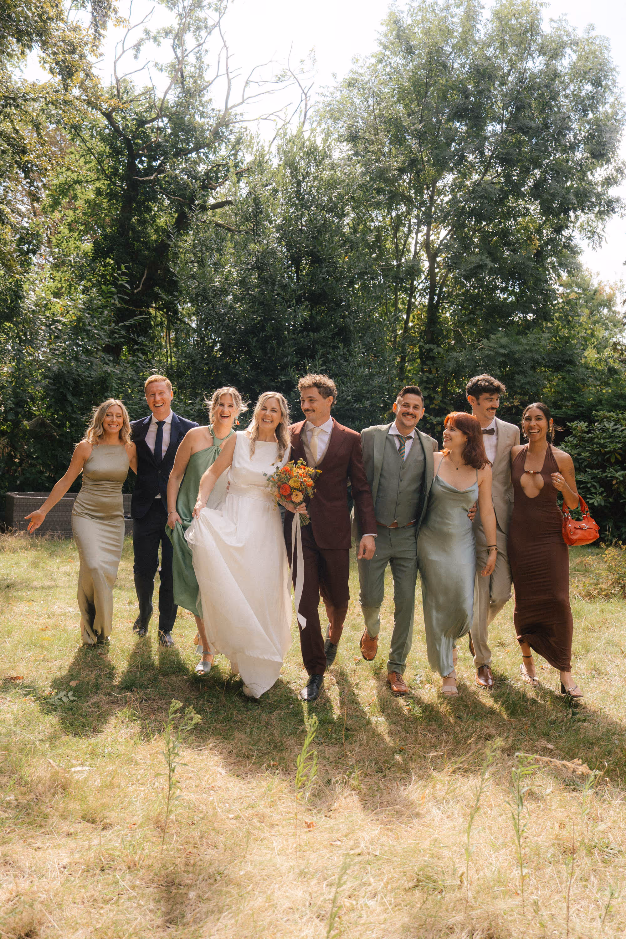 A joyful wedding party walking on grass with trees in the background, featuring a bride in a white dress holding a bouquet and friends in formal attire.