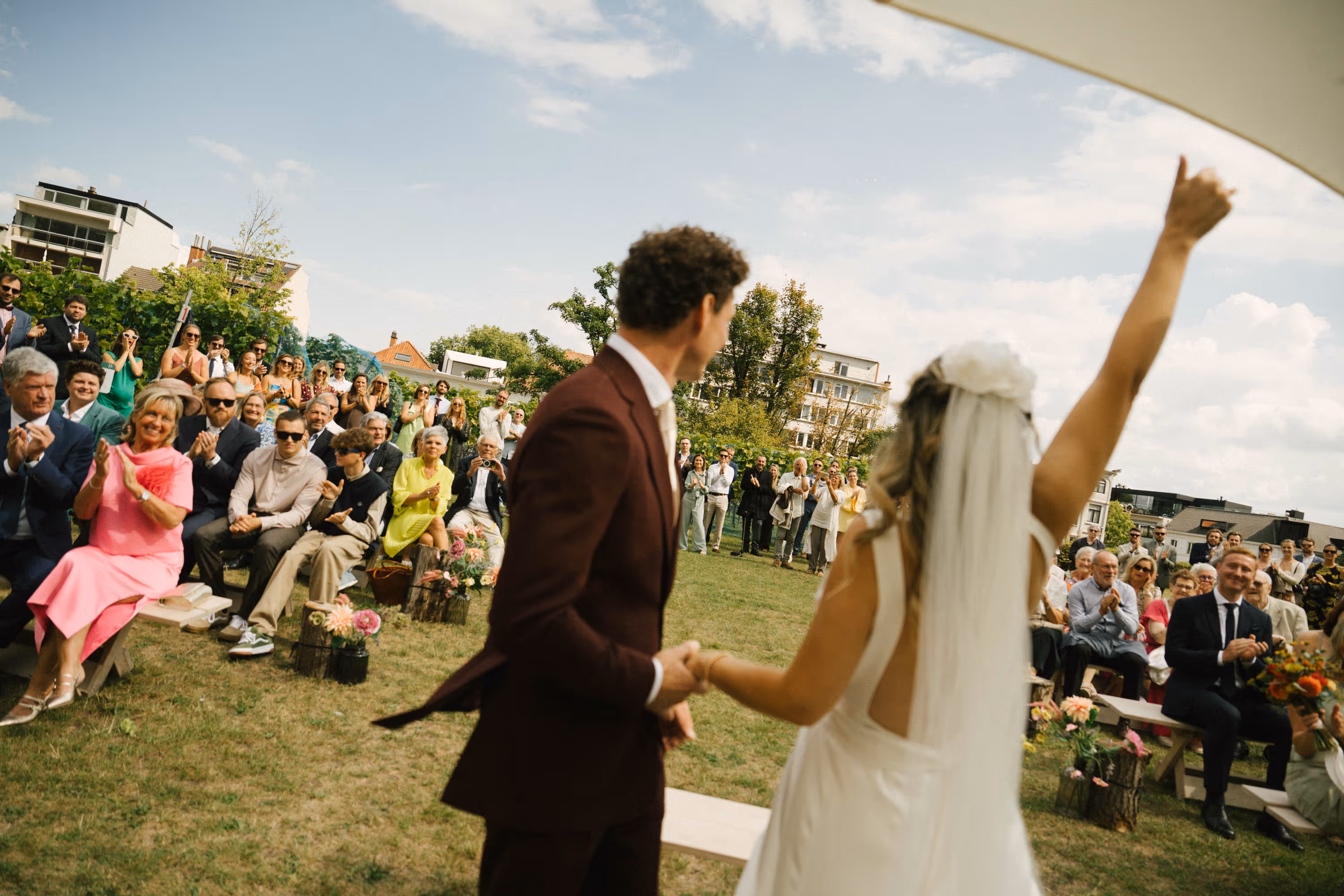 Bride in white dress raising arm with groom in dark suit holding hands, as guests seated outdoors applaud at wedding ceremony.