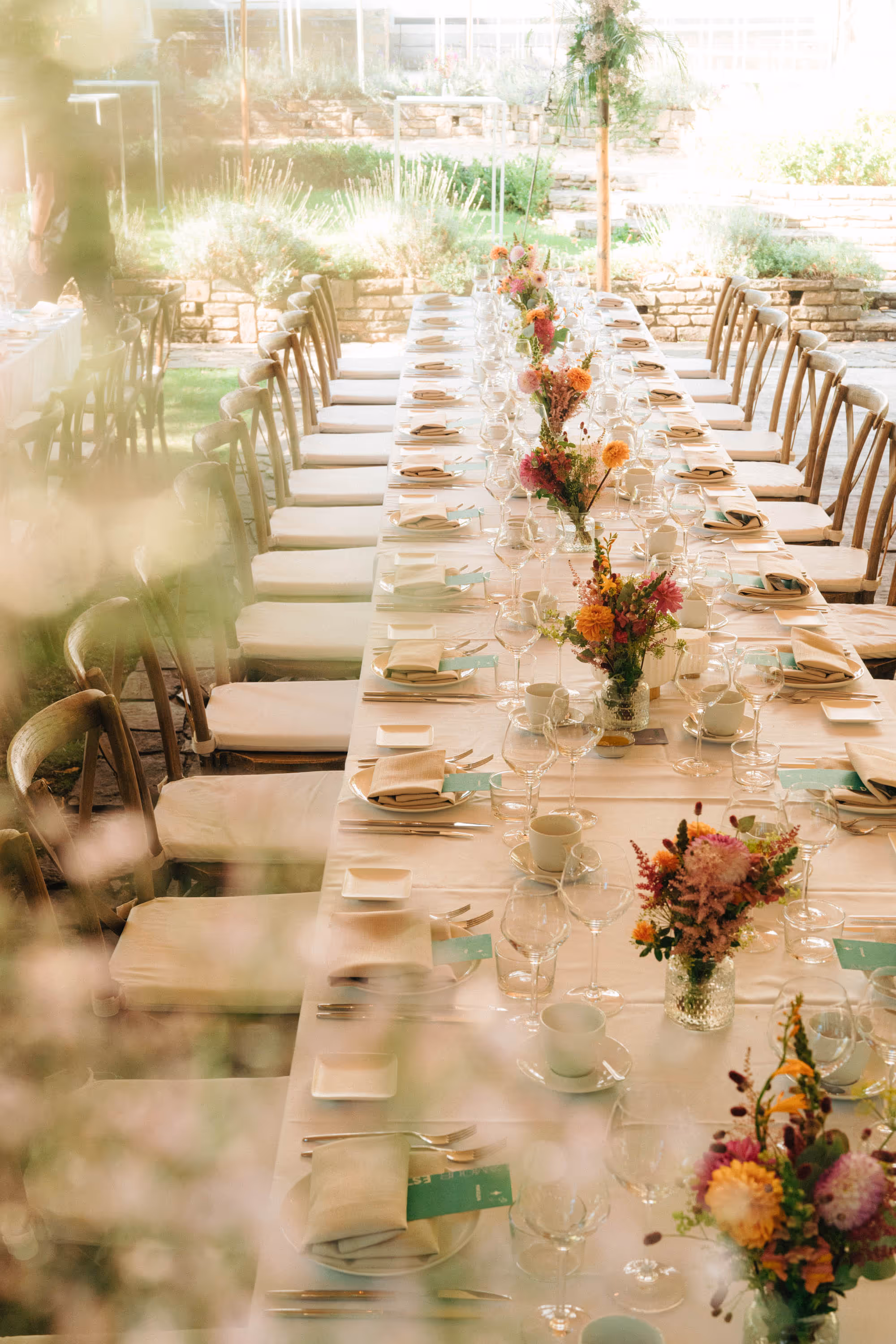 Long table set for an outdoor event with white tablecloth, floral centerpieces, glassware, and beige cushioned wooden chairs.