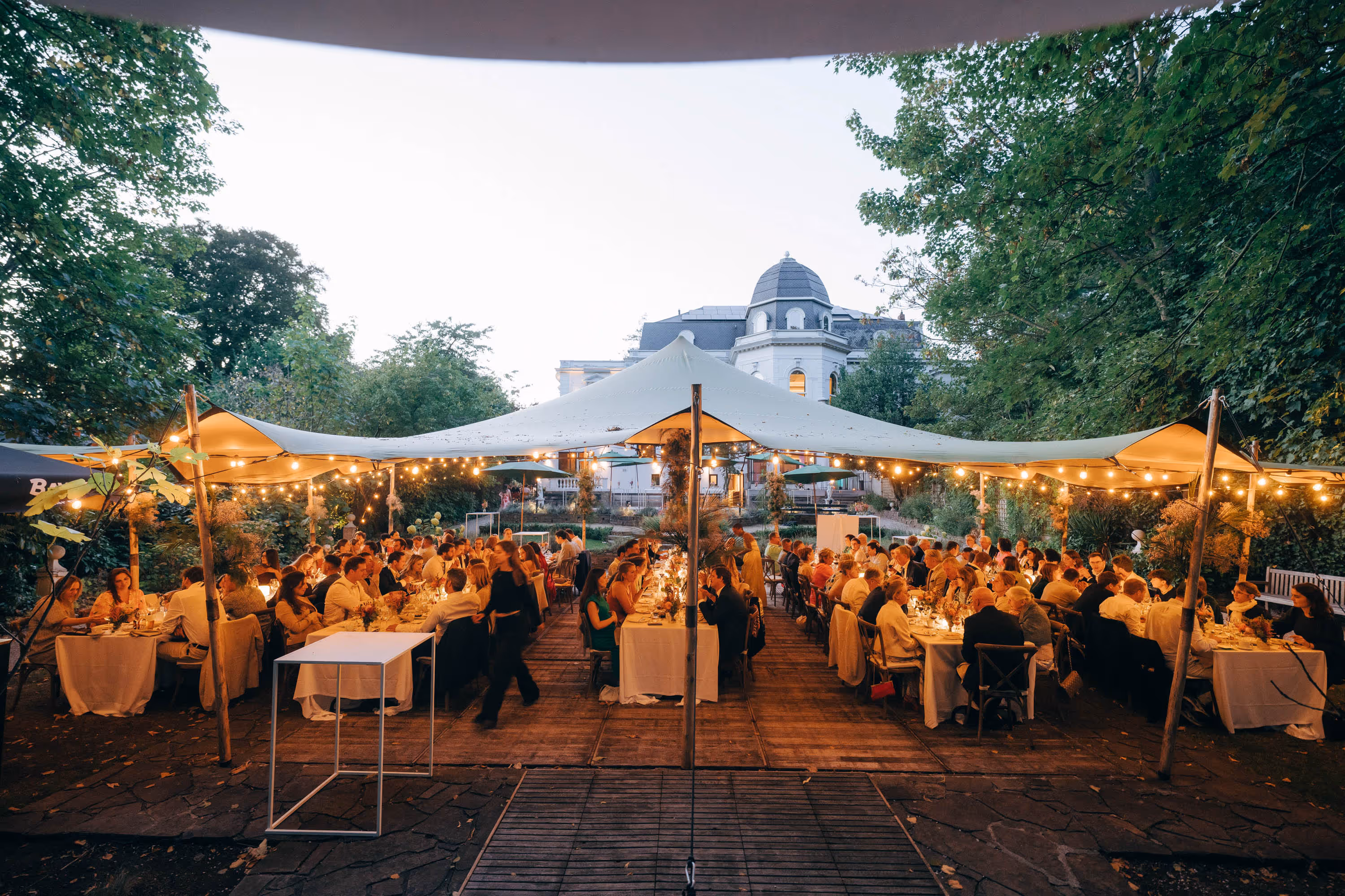 Outdoor evening dinner party under a large canopy with string lights and guests seated at long tables.