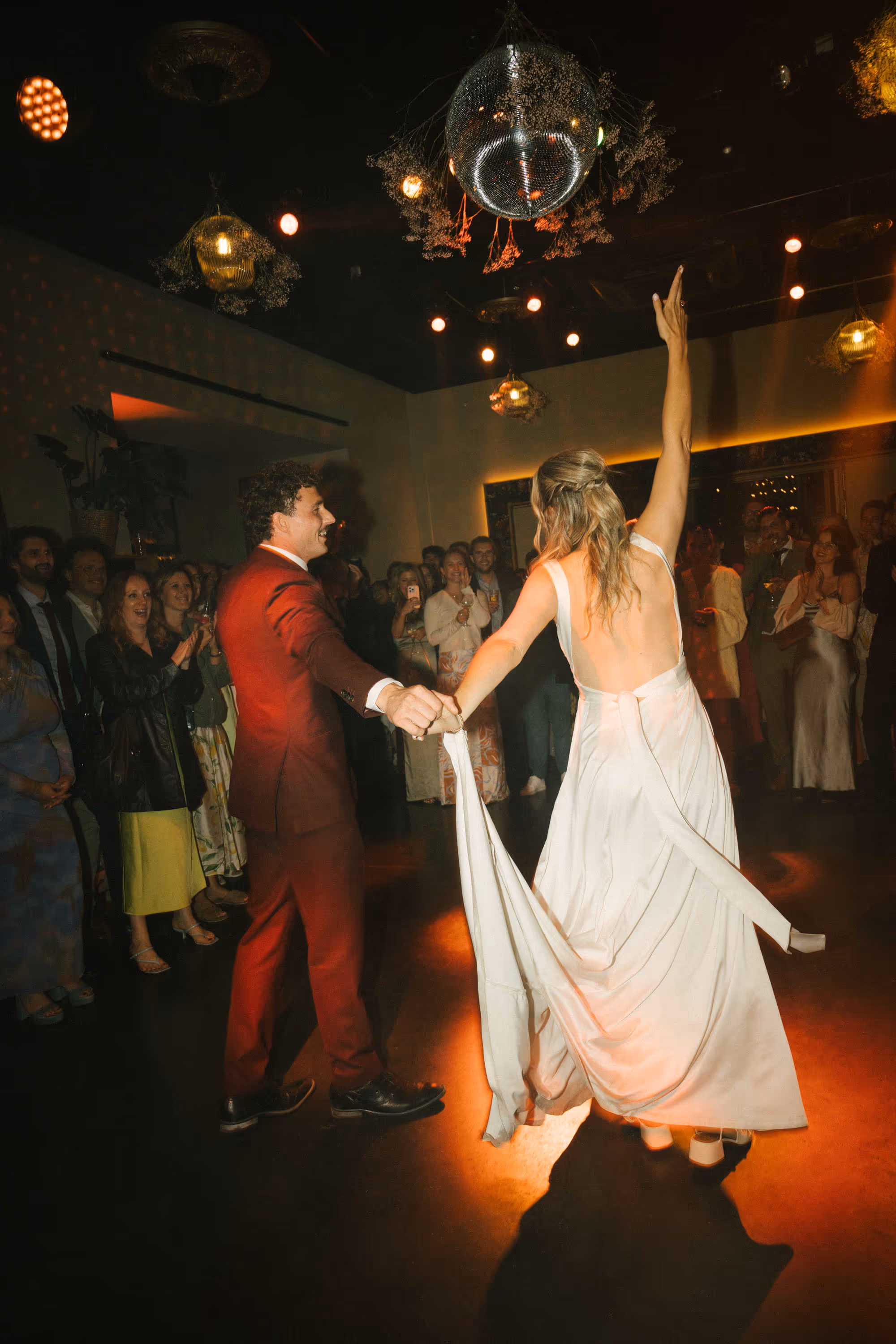 Bride in a white open-back dress and groom in a red suit dancing together surrounded by applauding guests under a disco ball.