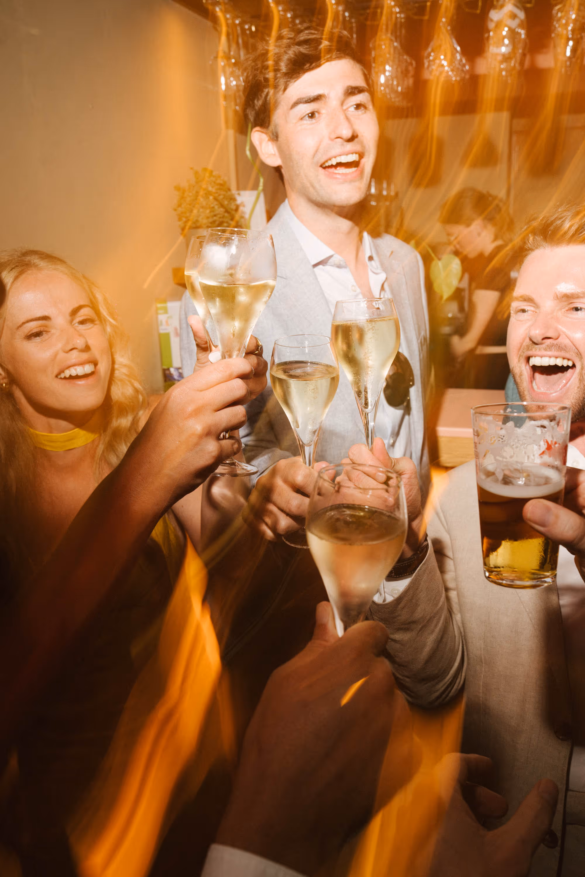 Group of friends smiling and toasting with champagne and beer glasses in a warmly lit indoor setting.