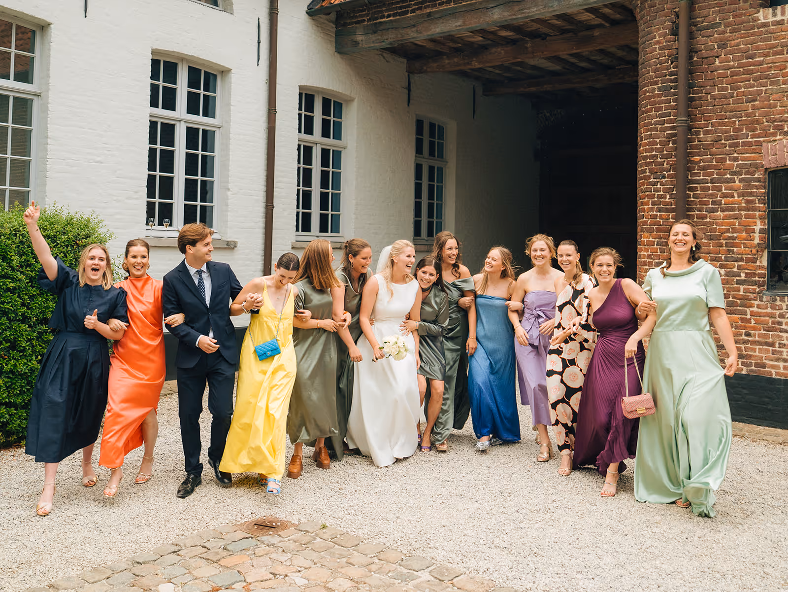 Group of people in colorful formal dresses and a suit walking arm-in-arm outdoors, smiling and laughing.