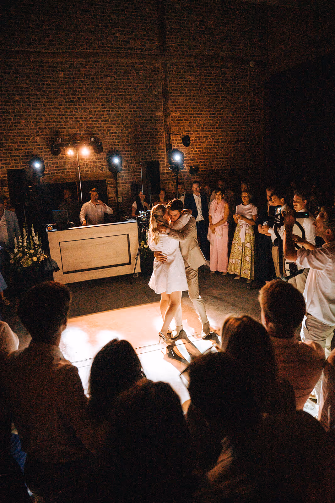 Couple embraces closely while dancing on a lighted floor surrounded by watching guests in a dimly lit venue with brick walls.
