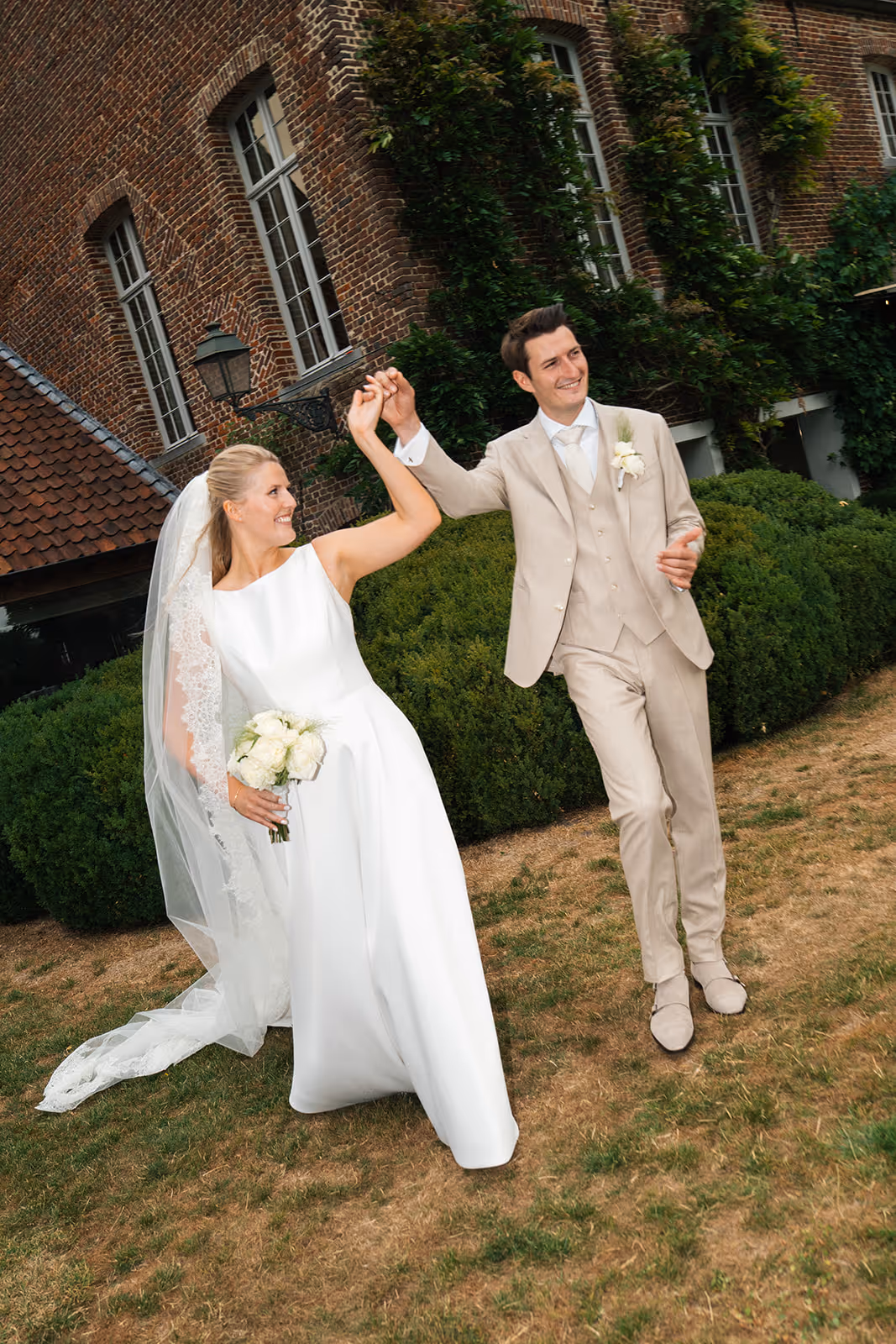 Bride in white dress and veil holding a bouquet of white flowers dancing with groom in beige suit outside in front of a brick building and greenery.