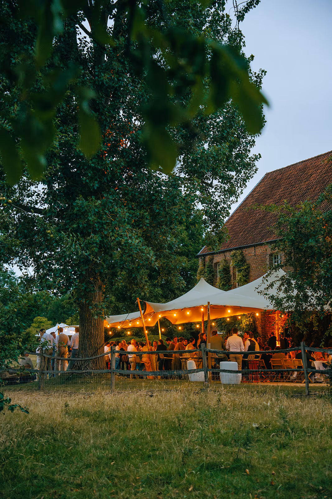 Outdoor evening gathering under a lit canopy tent near a brick building surrounded by trees and a wooden fence.