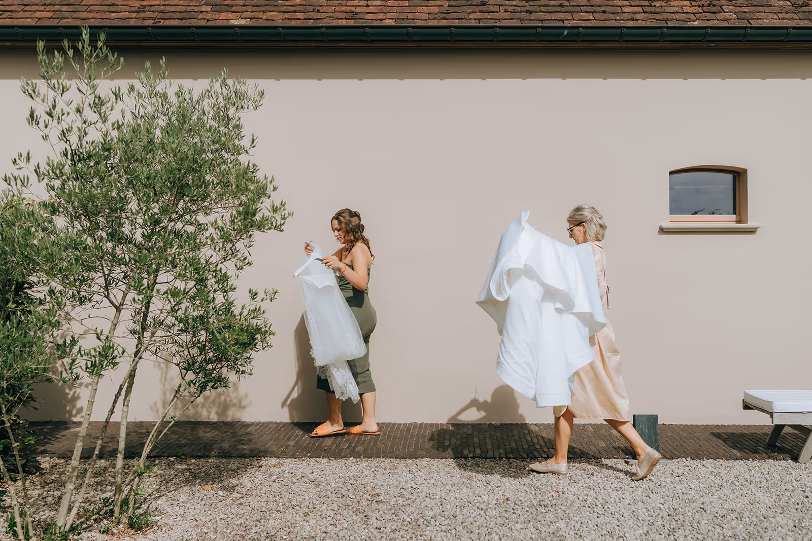 Two women outdoors against a beige wall, one holding a white dress and the other carrying a large white garment.