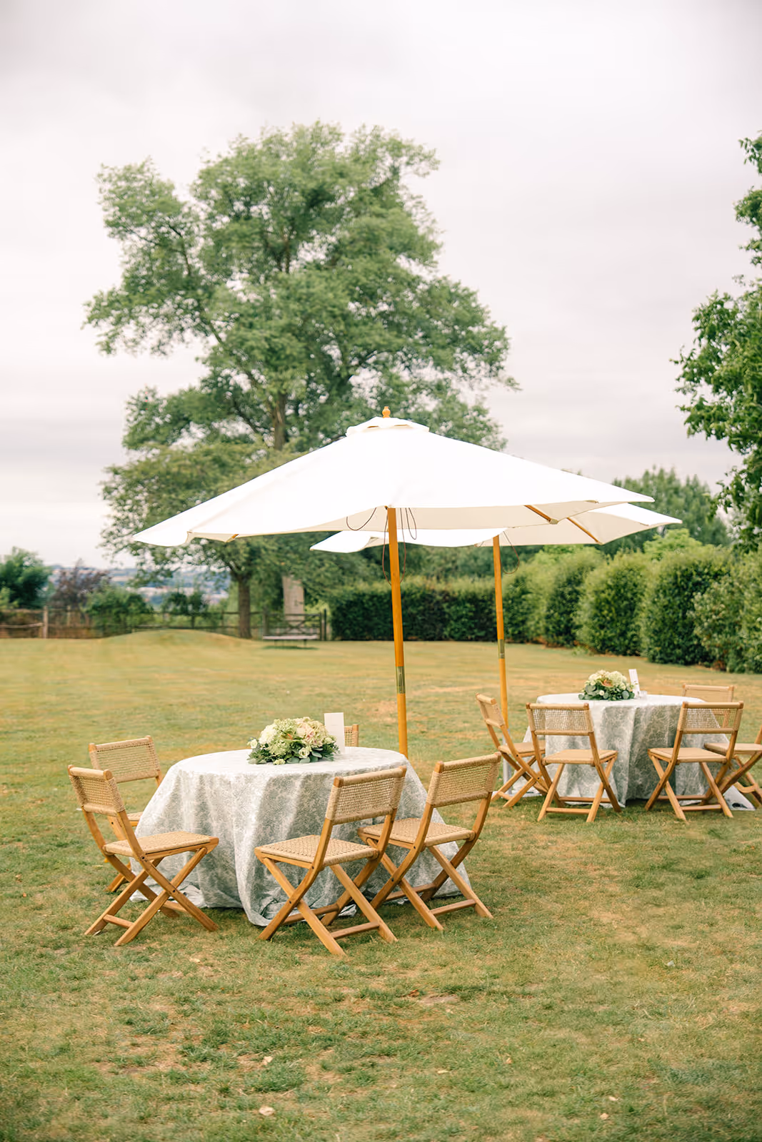 Outdoor garden setting with two round tables covered in light grey tablecloths, surrounded by wooden chairs and shaded by large white umbrellas.