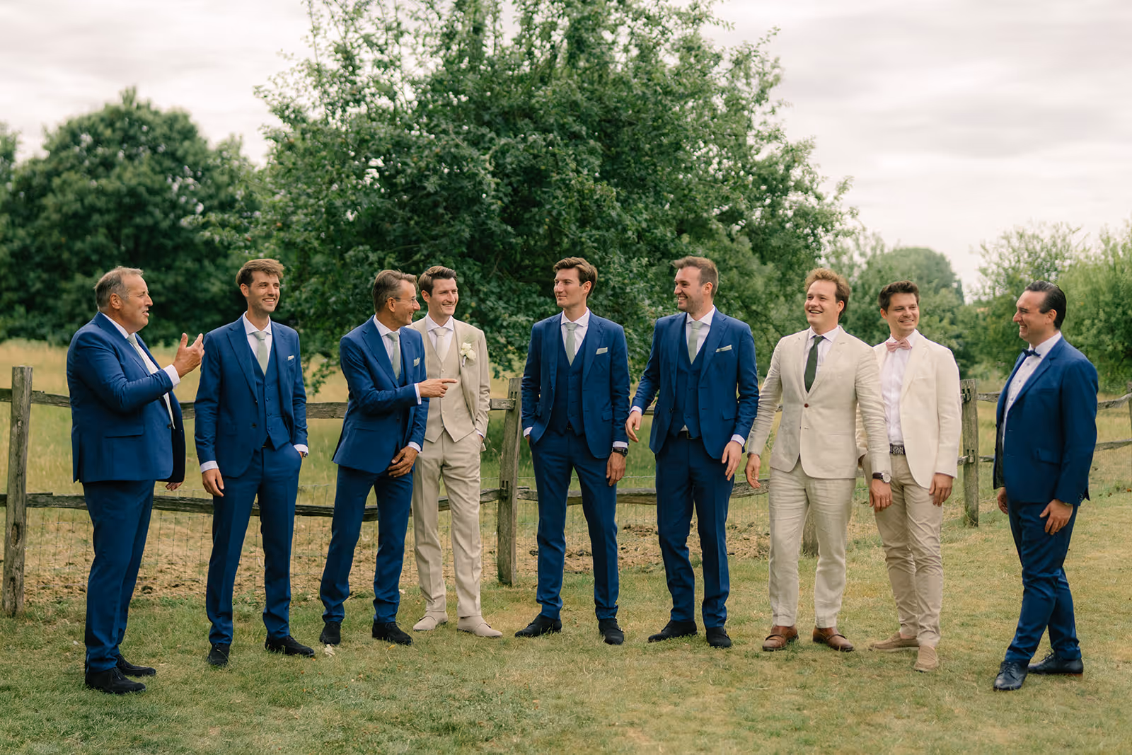Group of nine men in suits, with five wearing blue and four in beige or cream, standing outdoors near a wooden fence and trees.
