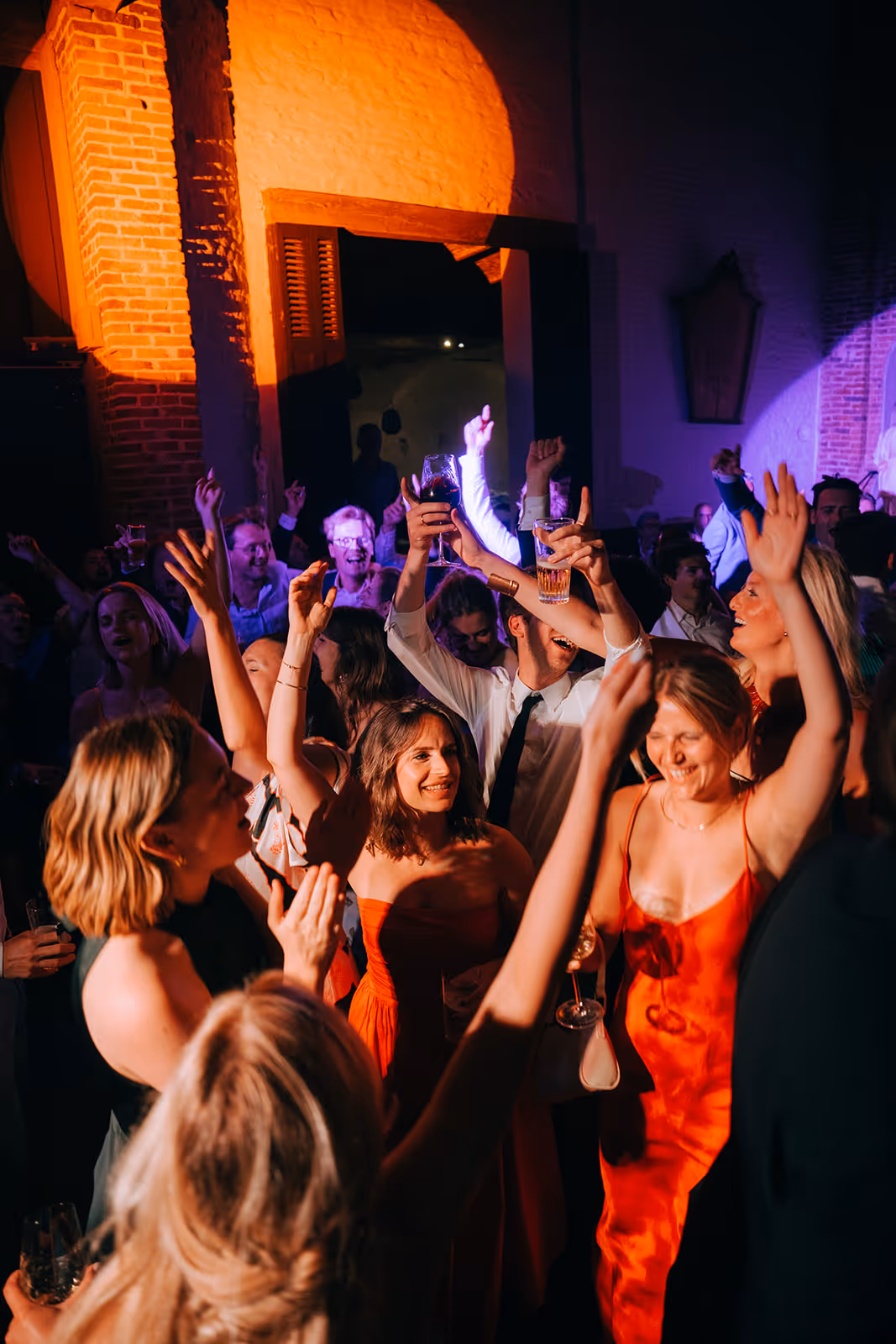 Group of people dancing and celebrating indoors with drinks raised, illuminated by warm and purple lighting.