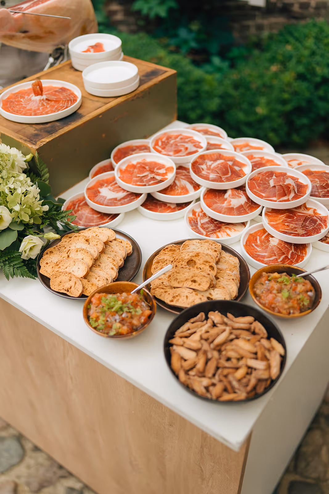 Table with plates of sliced cured ham, bowls of salsa and nuts, and plates of toasted bread slices.