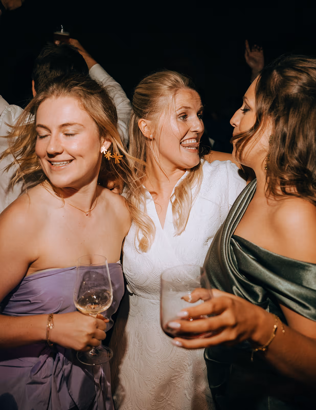 Three women smiling and enjoying a celebration, holding drinks and dressed in elegant evening wear.