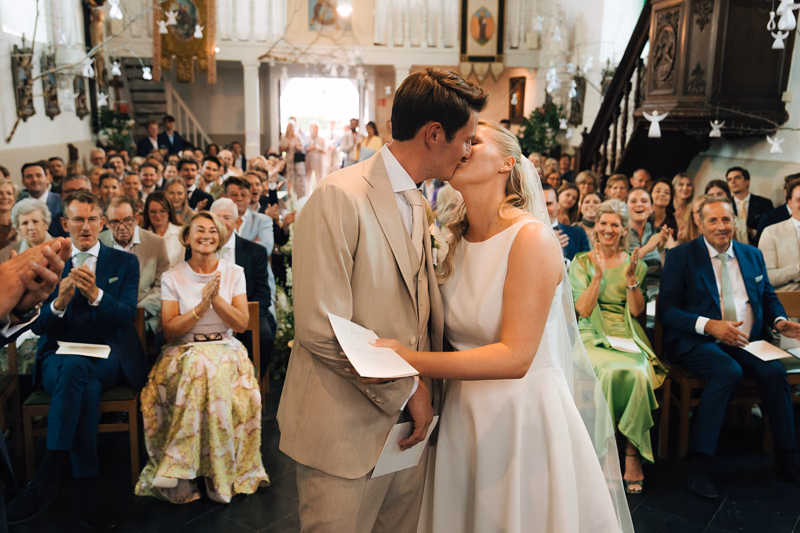 Bride and groom kissing in church during wedding ceremony with guests clapping and smiling in the background.