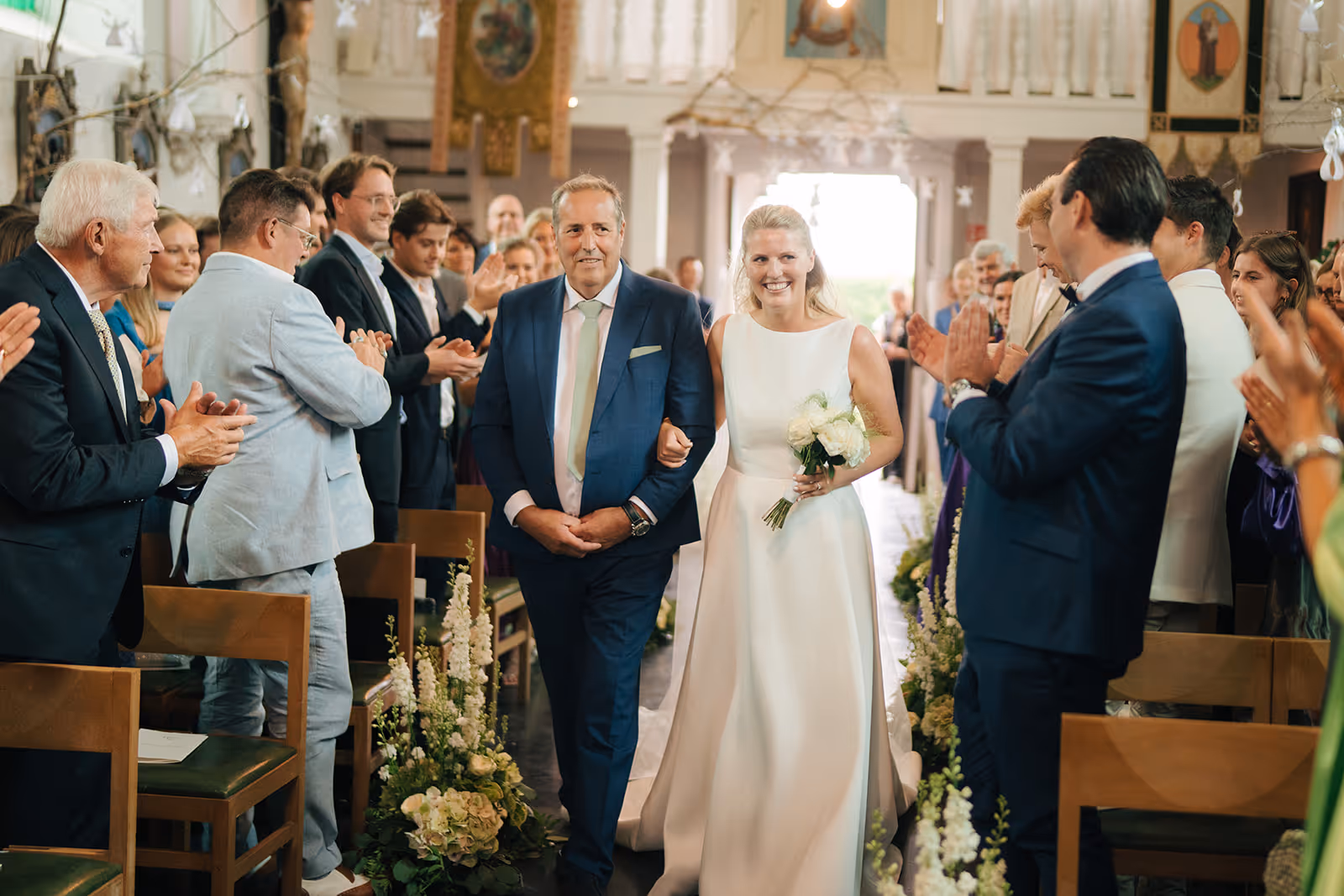 Bride in a white dress holding a bouquet walking arm-in-arm with a man in a blue suit down a flower-lined aisle while guests applaud.