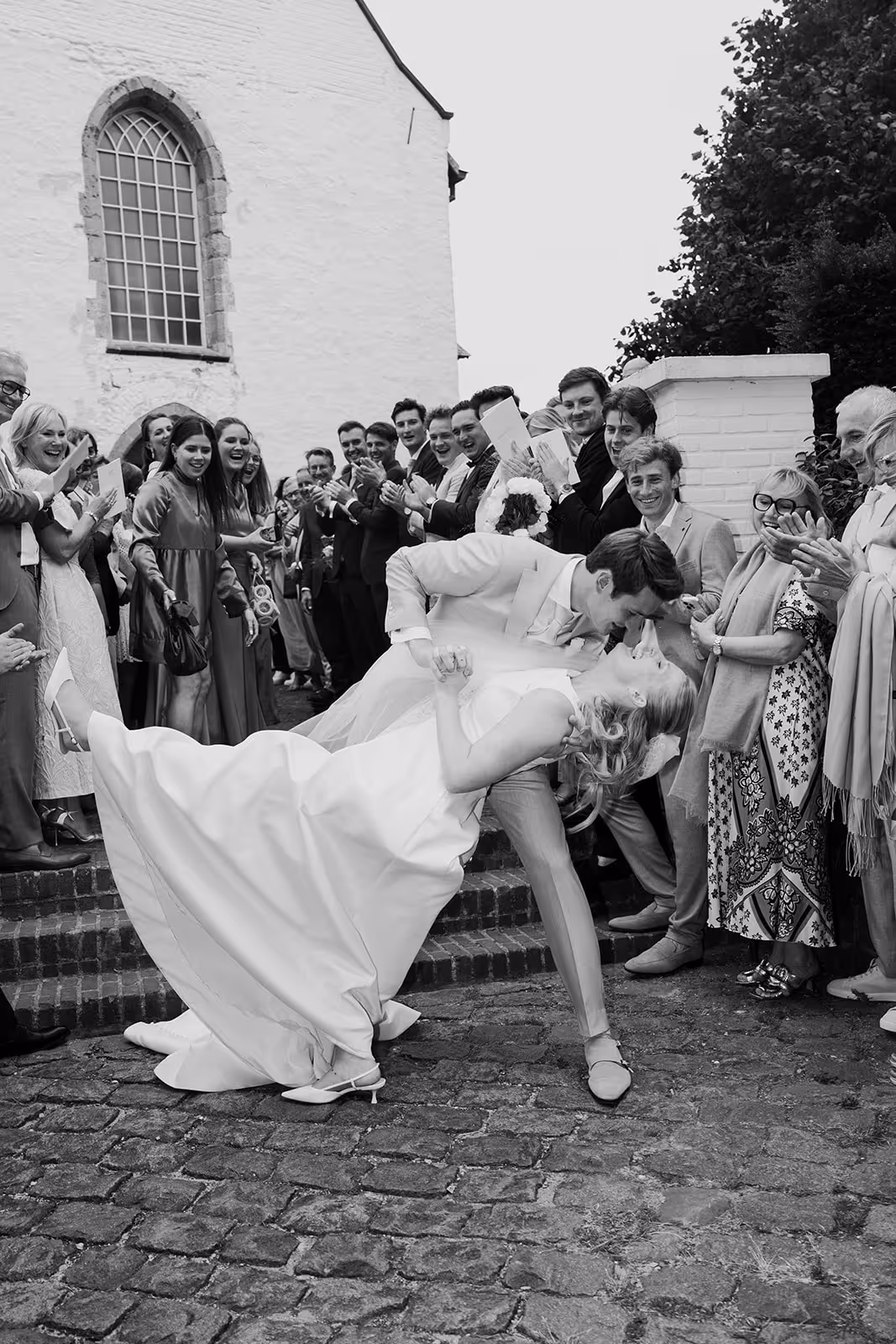 Bride and groom sharing a kiss in a dance dip while guests applaud outside a church.
