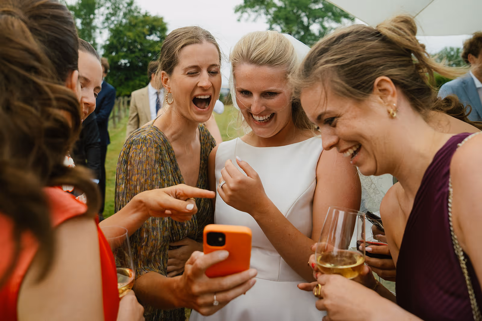 Group of women, including a bride in a white dress, laughing and looking at a phone at an outdoor event.