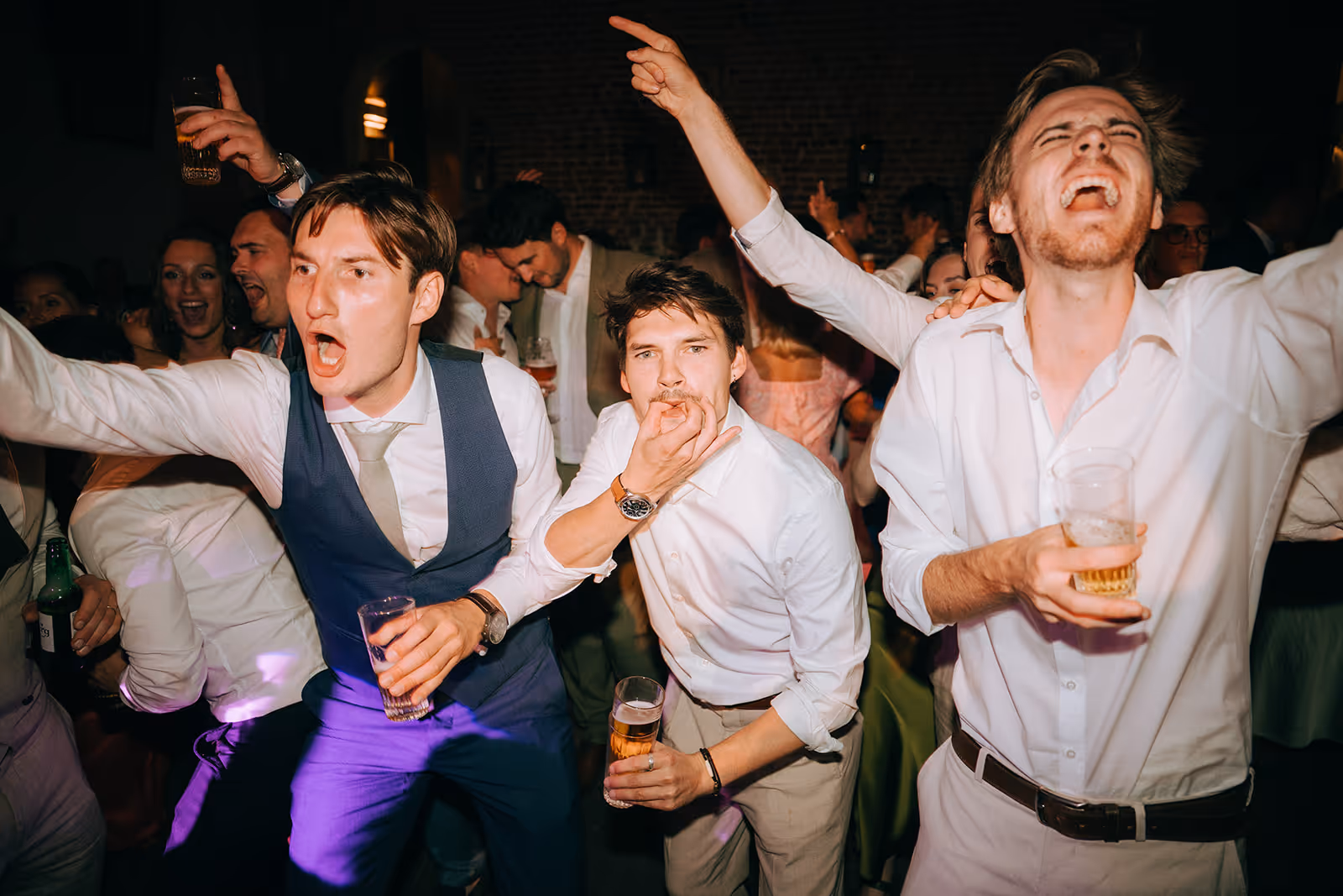 Three young men at a party enthusiastically singing or shouting with drinks in their hands.