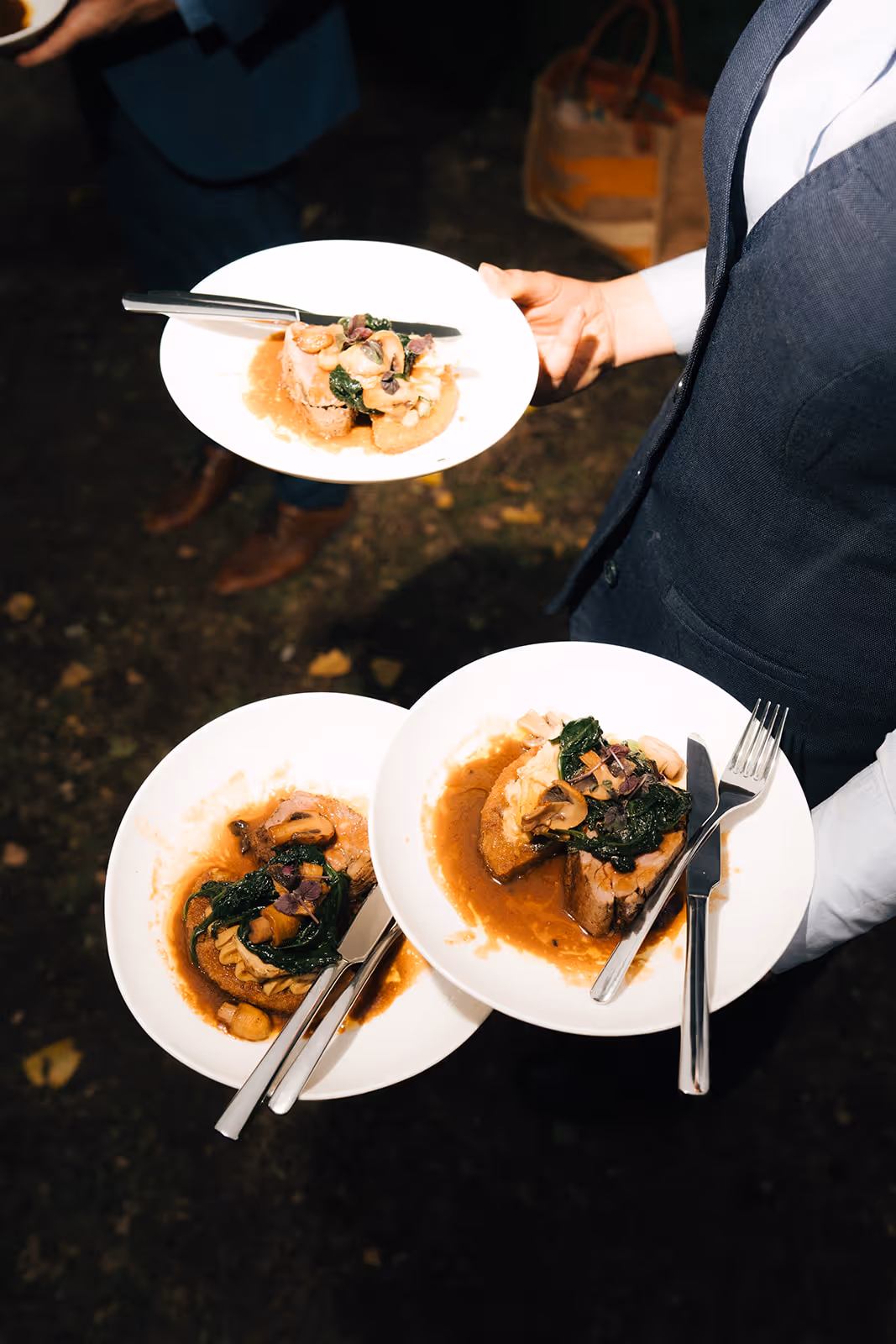 Waiter holding three white plates with gourmet dishes featuring meat, sautéed mushrooms, leafy greens, and brown sauce.