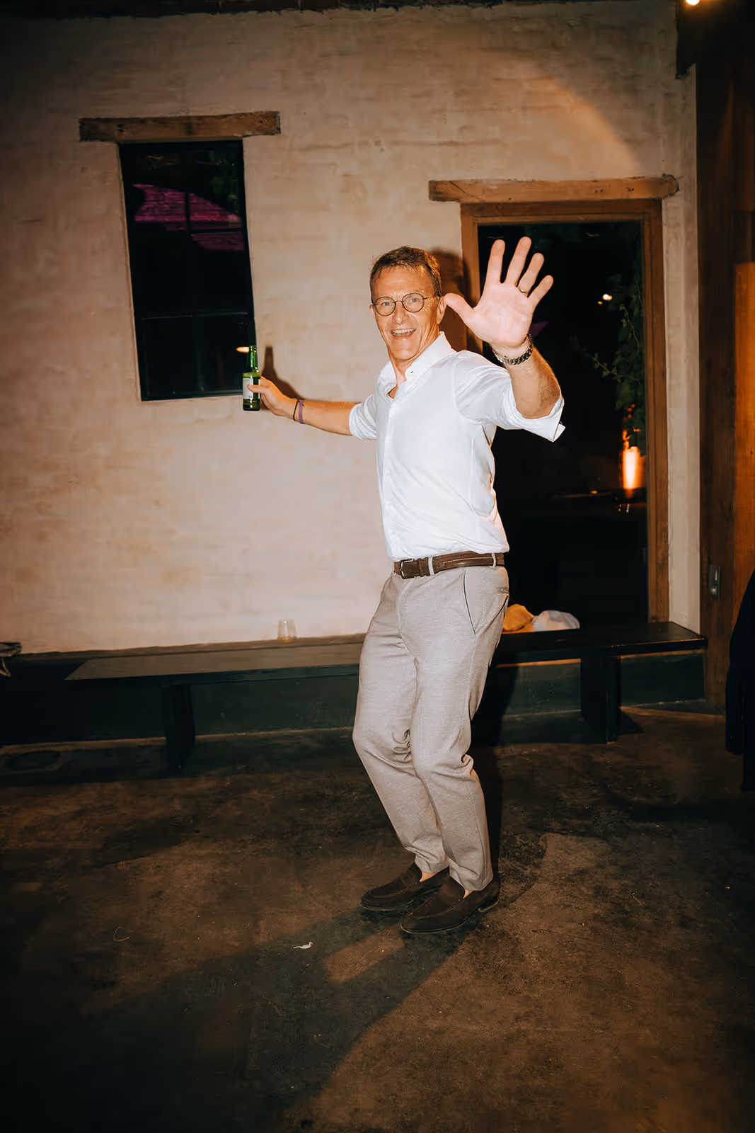 Smiling man wearing glasses and a white shirt, holding a bottle and waving with one hand indoors.