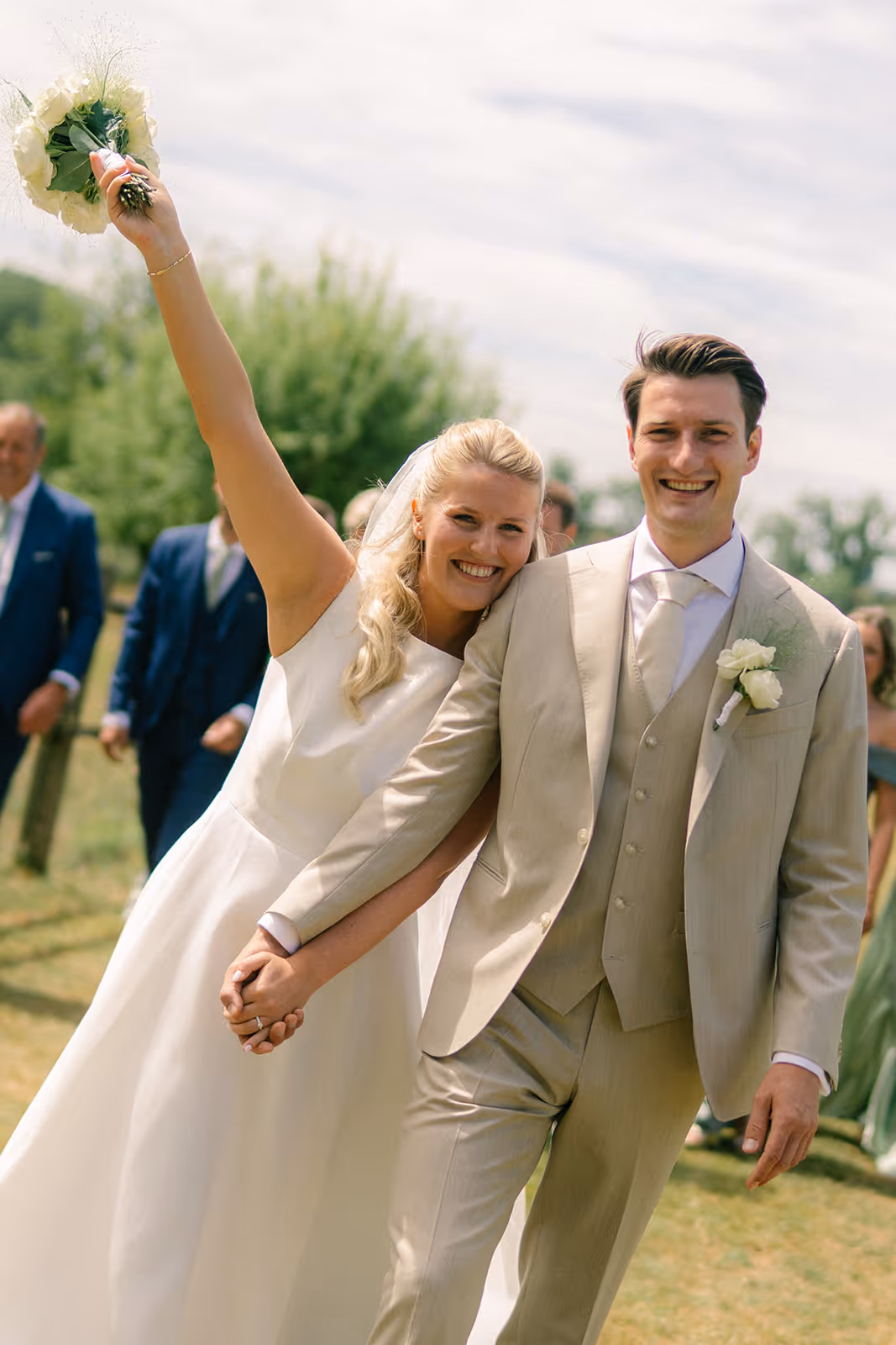 Smiling bride and groom holding hands outdoors, bride raising bouquet in celebration.