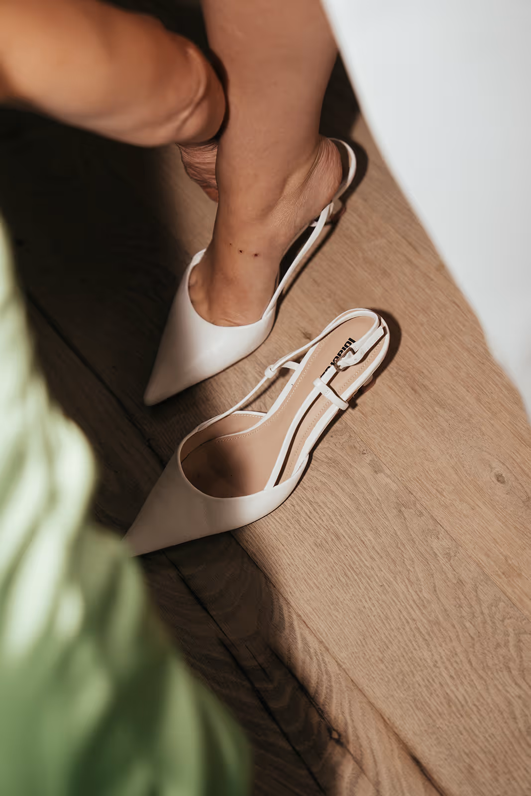 Person wearing white pointed slingback heels on wooden floor with a second shoe nearby.