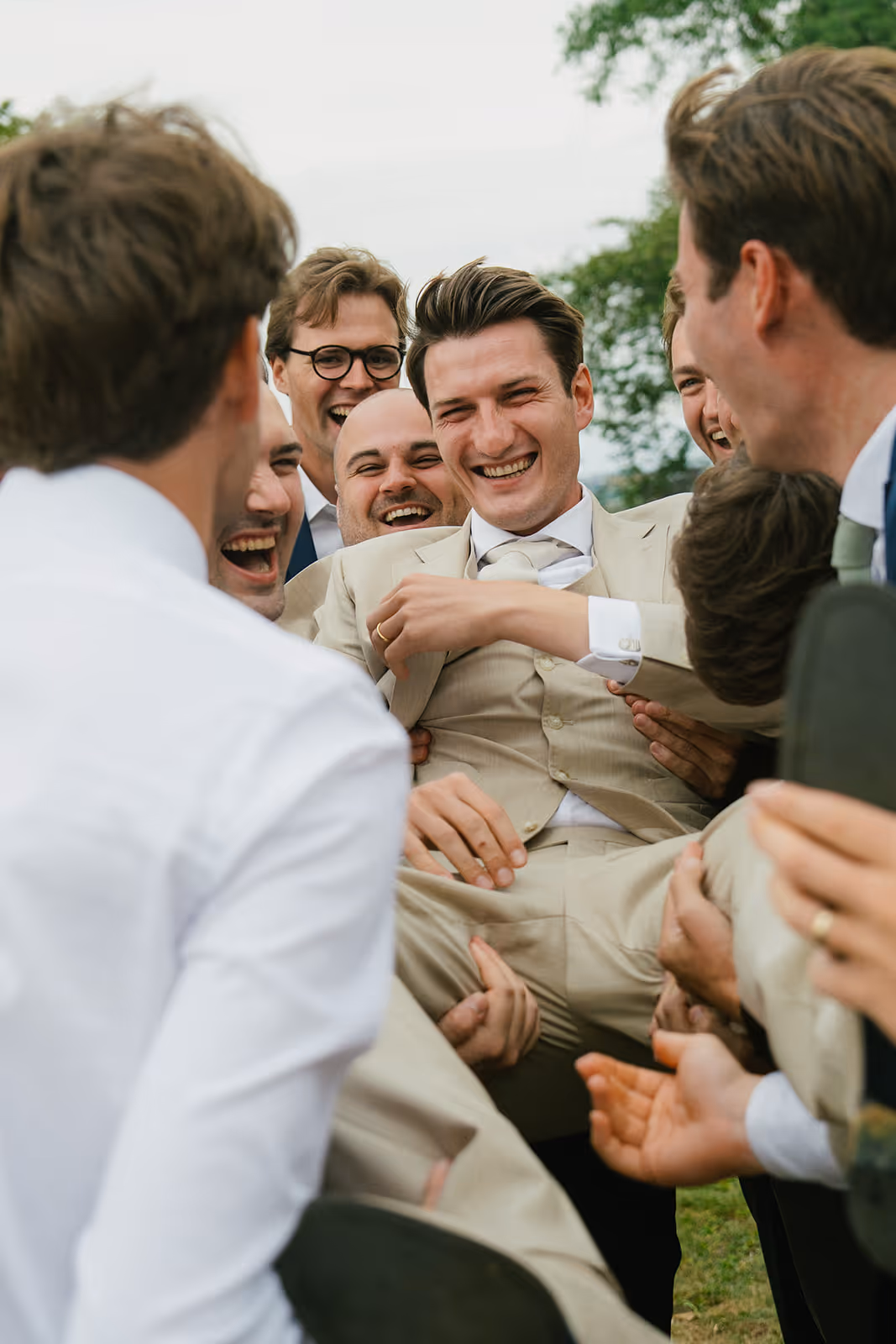 Group of men joyfully lifting a groom in a beige suit during an outdoor celebration.