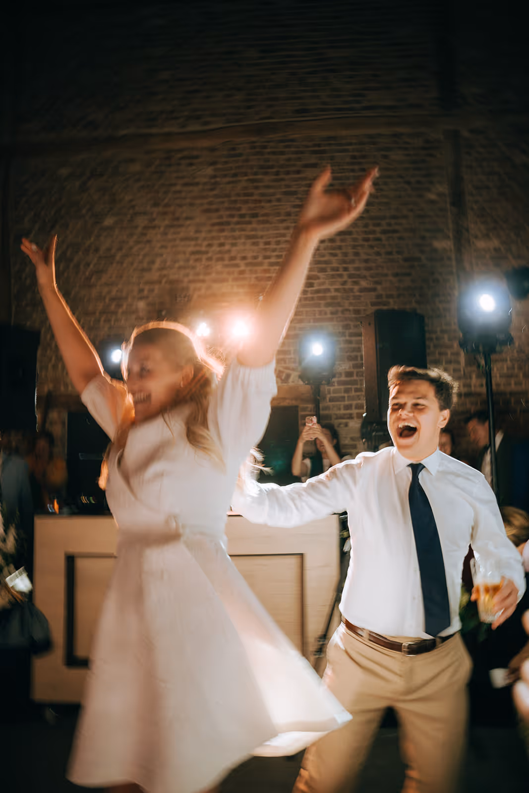 Man and woman dancing energetically at a party with lights and brick wall background.