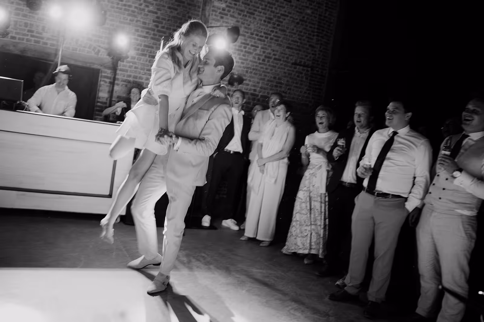 Black and white photo of a man lifting a woman while dancing, surrounded by smiling and clapping guests at a celebration indoors.