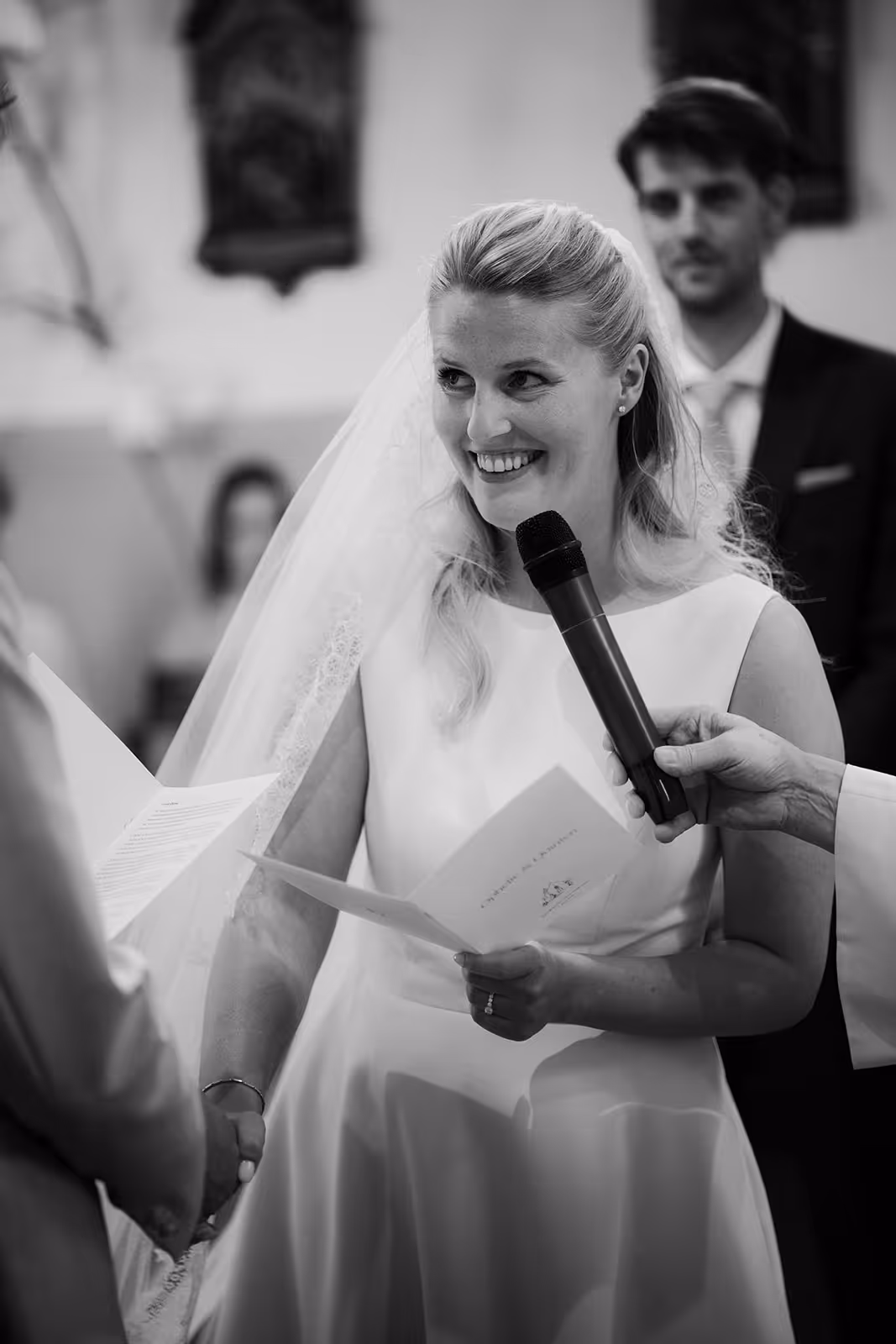 Smiling bride in a wedding dress holding a paper and microphone, holding hands with the groom during the ceremony.