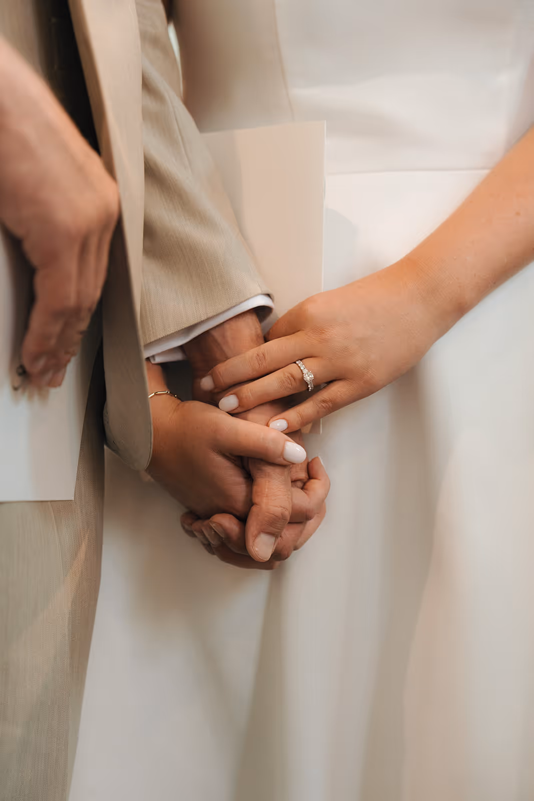 Close-up of a couple holding hands, the woman wearing a diamond engagement ring and both dressed in light-colored formal attire.
