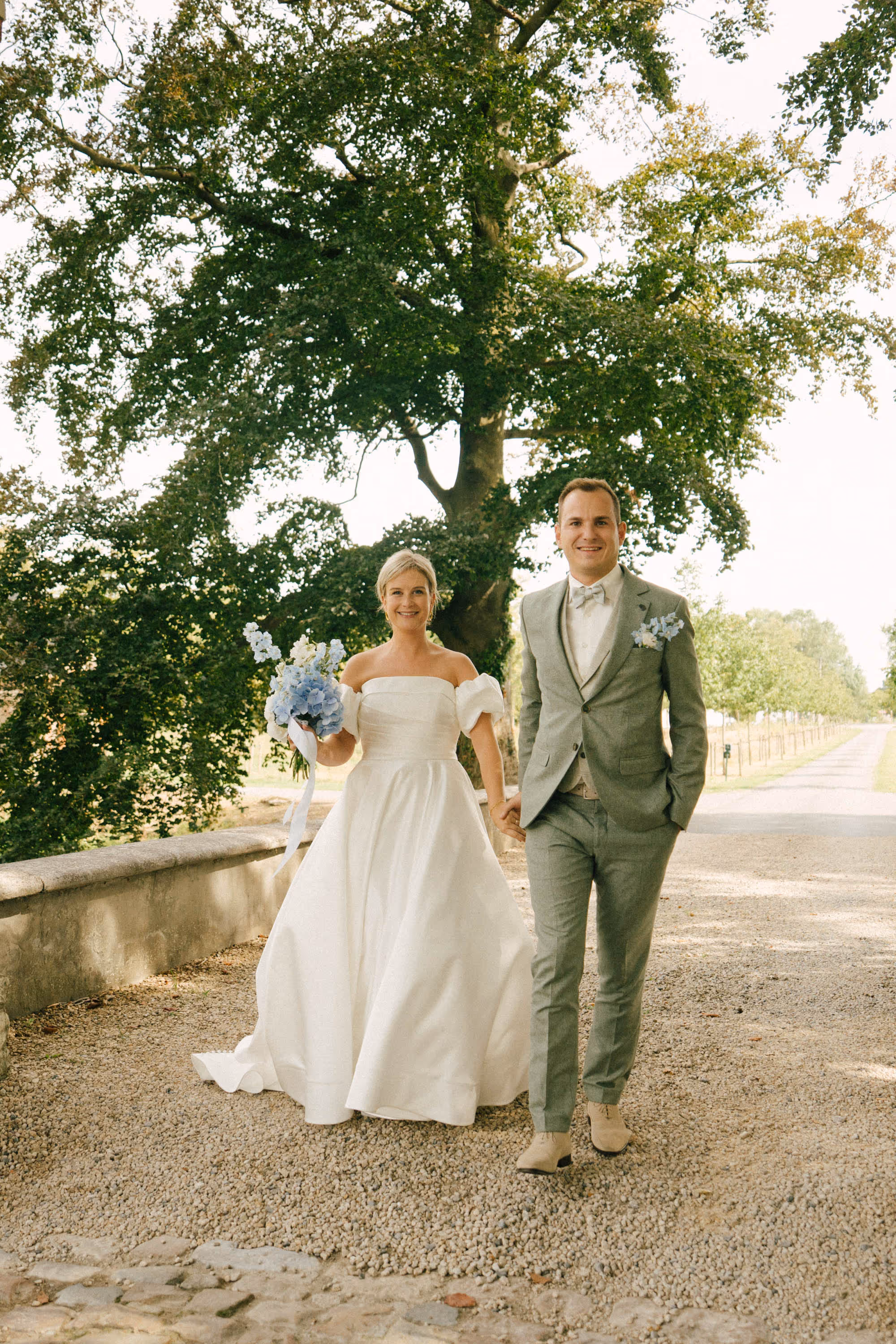 Bride in white dress holding a blue floral bouquet and groom in light gray suit walking hand in hand on a gravel path with trees in the background.