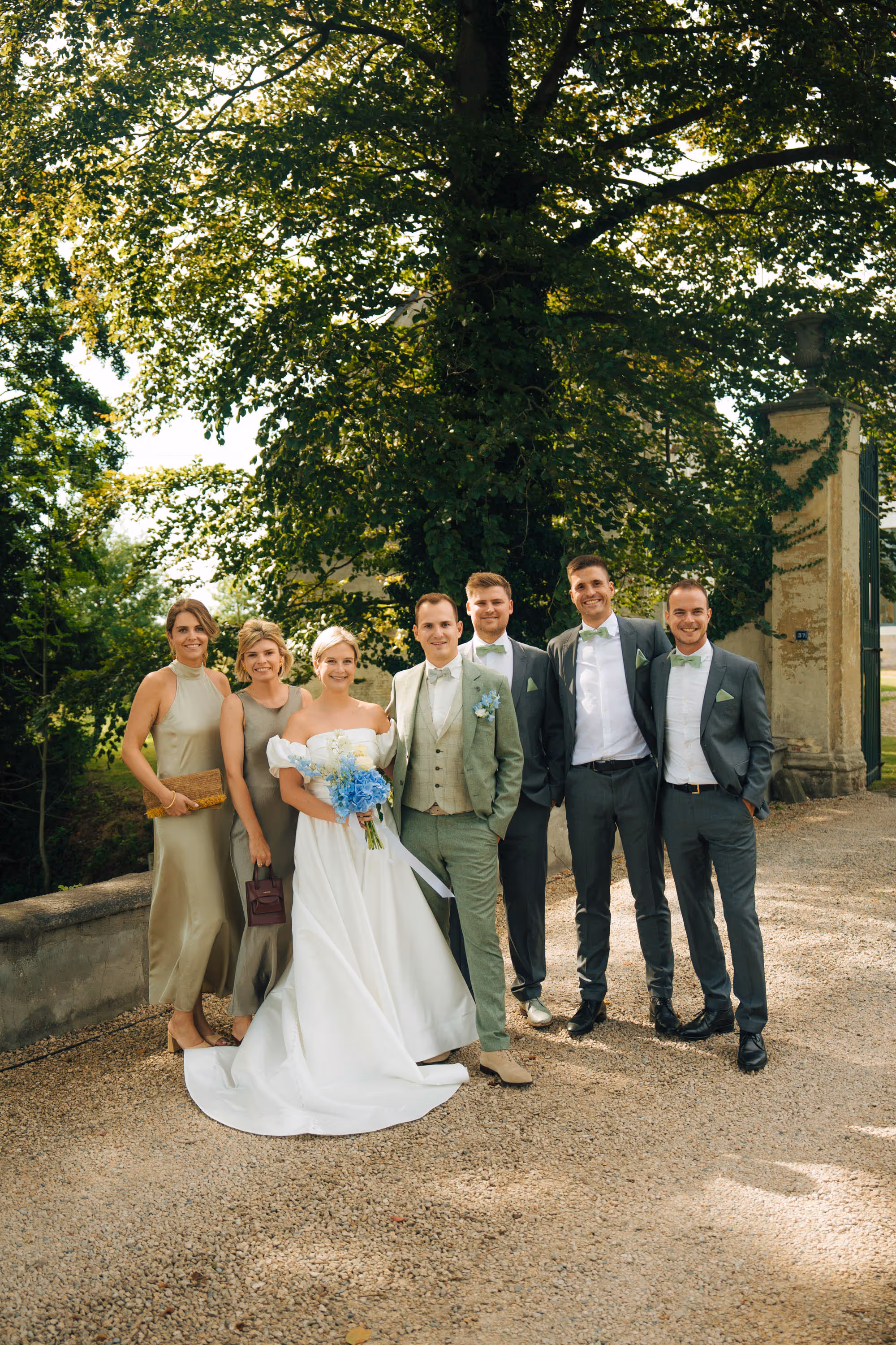 Bride in white dress holding blue bouquet with groom and wedding party dressed in coordinating green and gray attire outdoors.