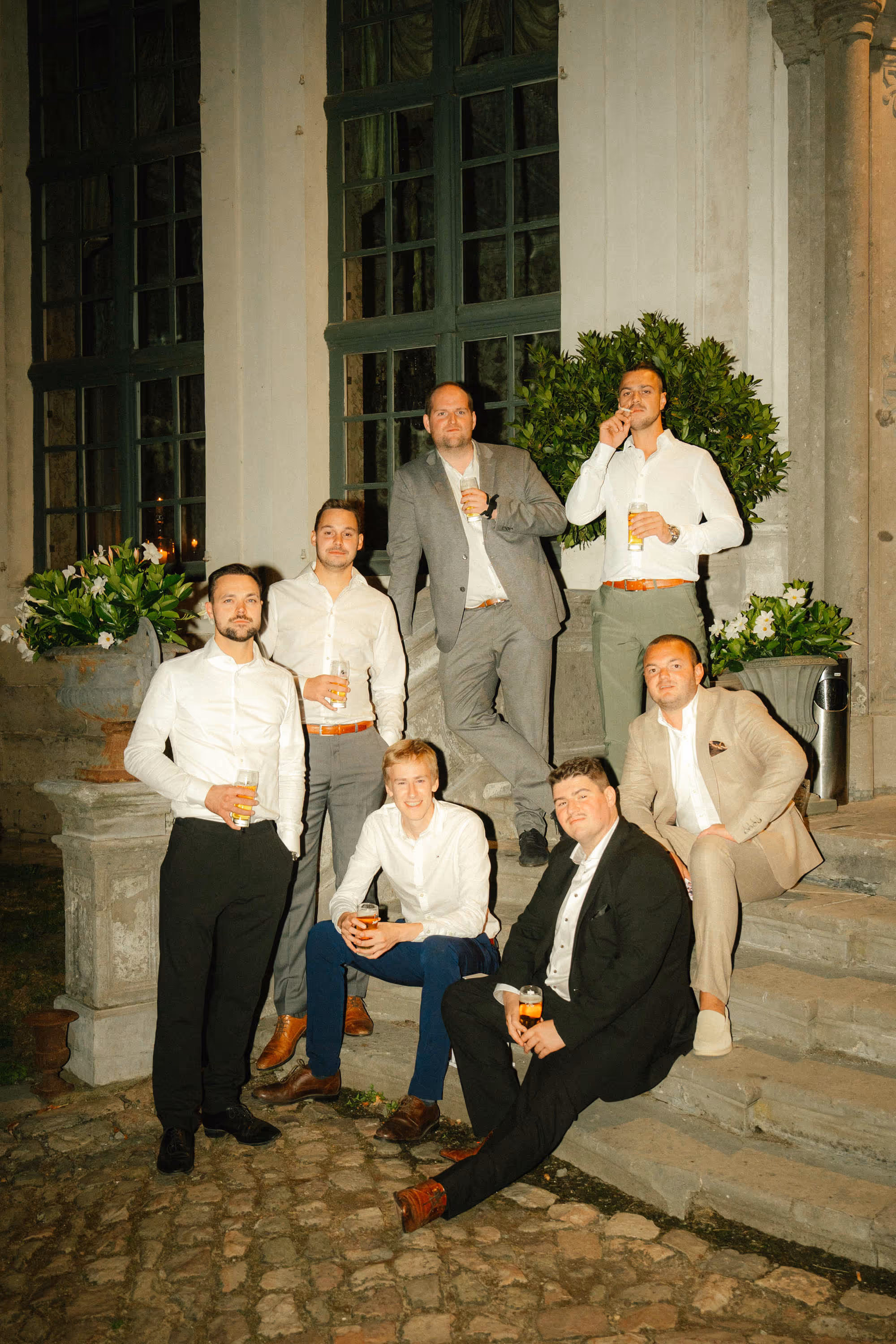 Seven men dressed in formal attire holding drinks, posing on stone steps outside a building at night.