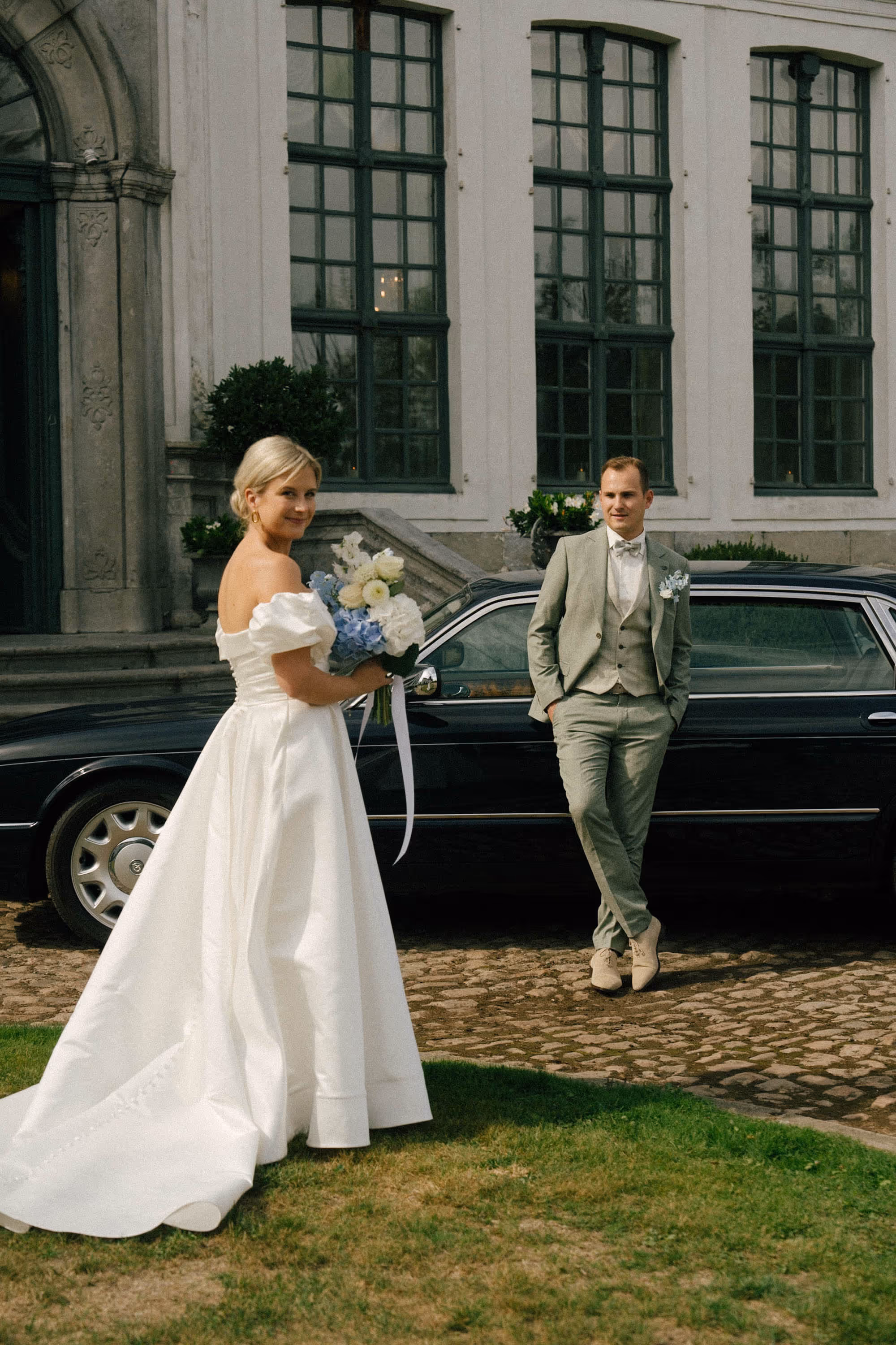 Bride in a white off-shoulder wedding dress holding a bouquet, and groom in a light gray suit leaning against a black car outside a building.