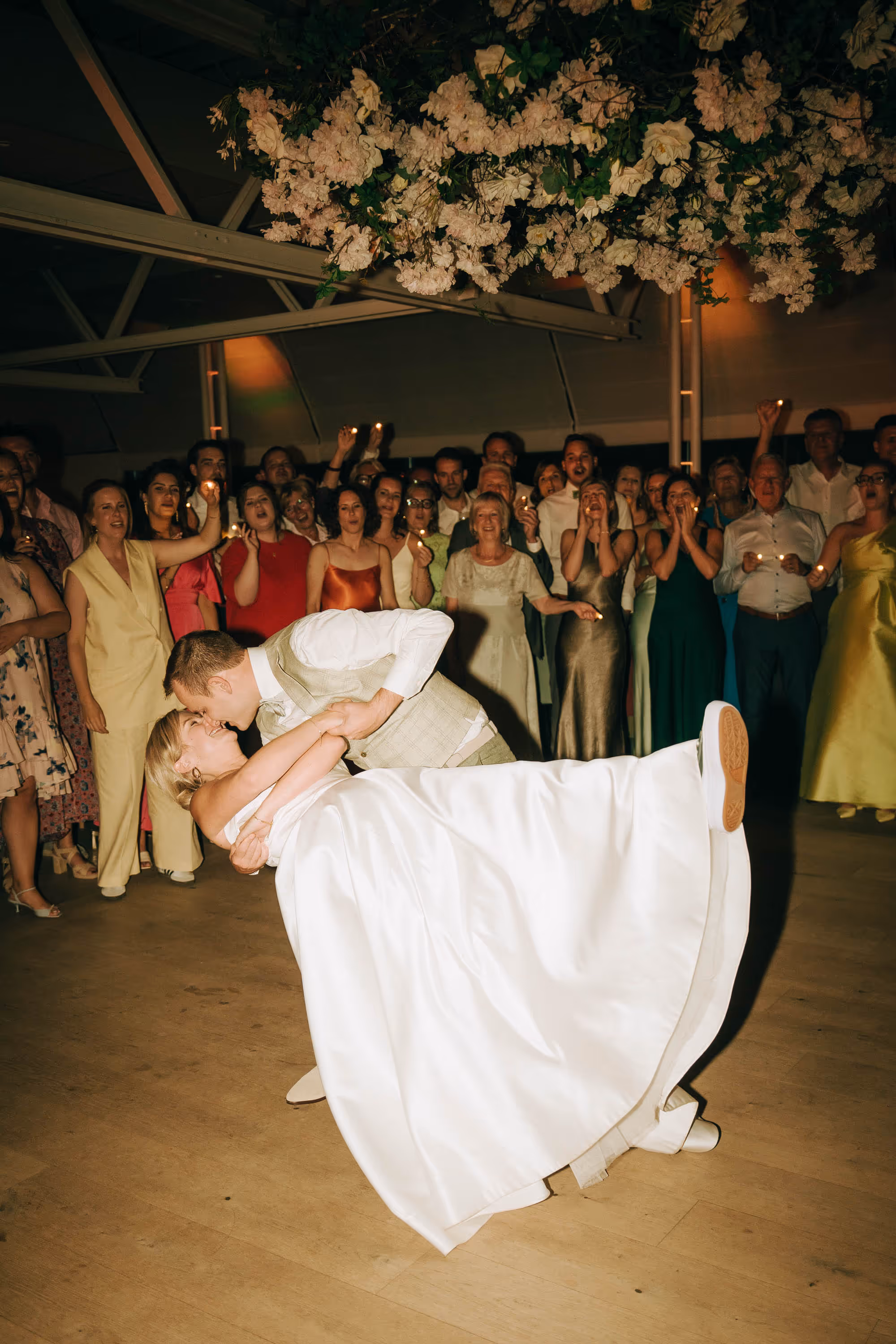 Bride in white dress dipped by groom wearing light-colored vest during wedding dance with crowd holding candles in the background.