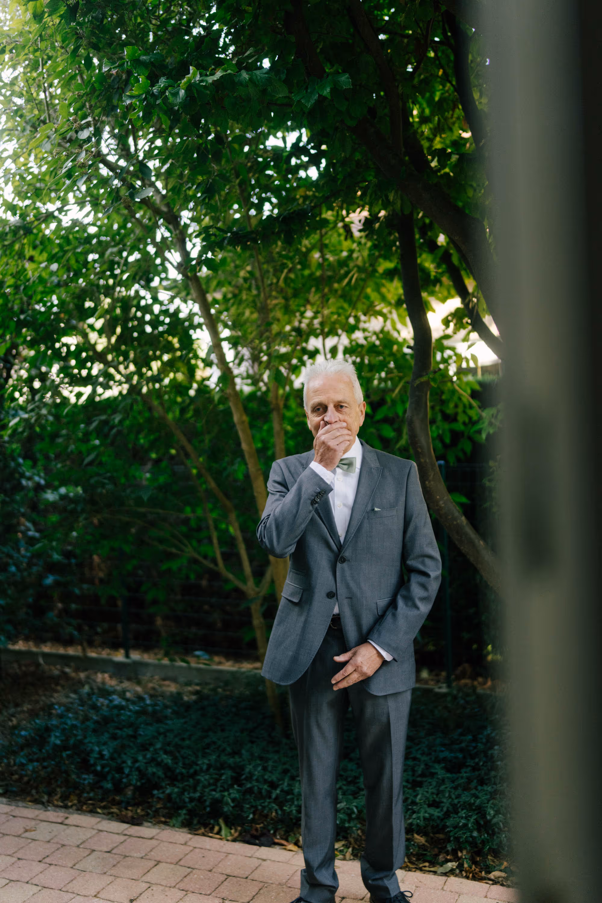 Elderly man in a gray suit standing outside under green trees with one hand covering his mouth.