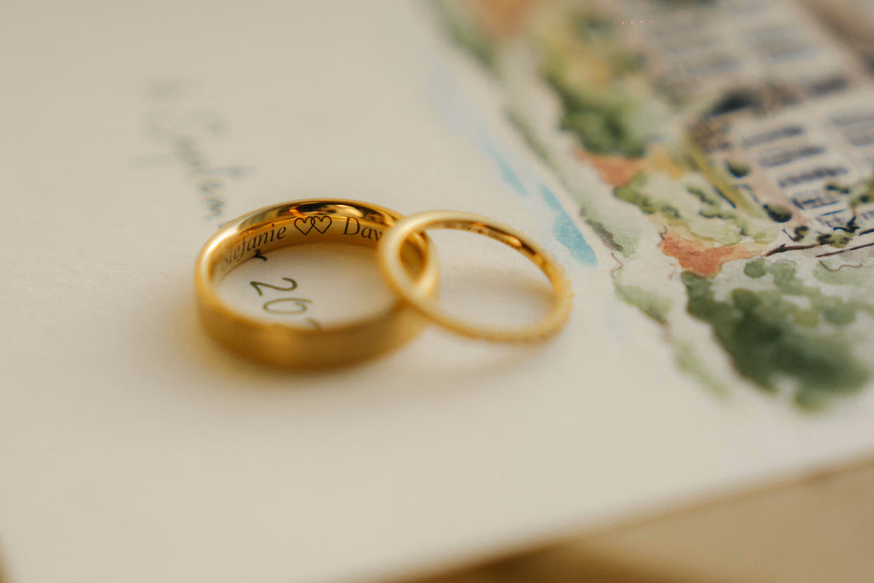 Two gold wedding rings on a paper with a watercolor illustration, one ring engraved with two hearts and partial names.