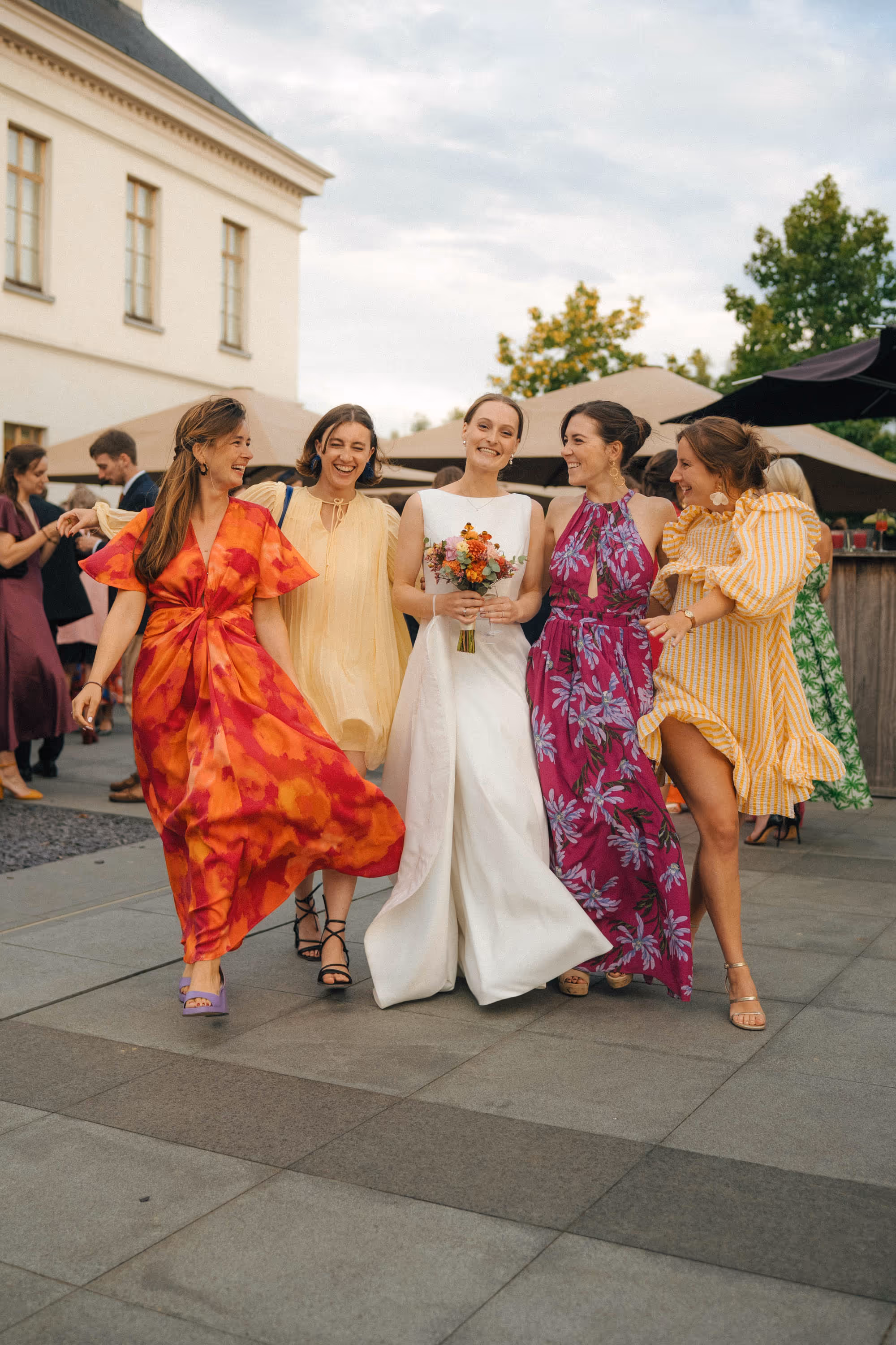 Bride in white dress holding bouquet walking arm-in-arm with four smiling women in colorful dresses at an outdoor event.