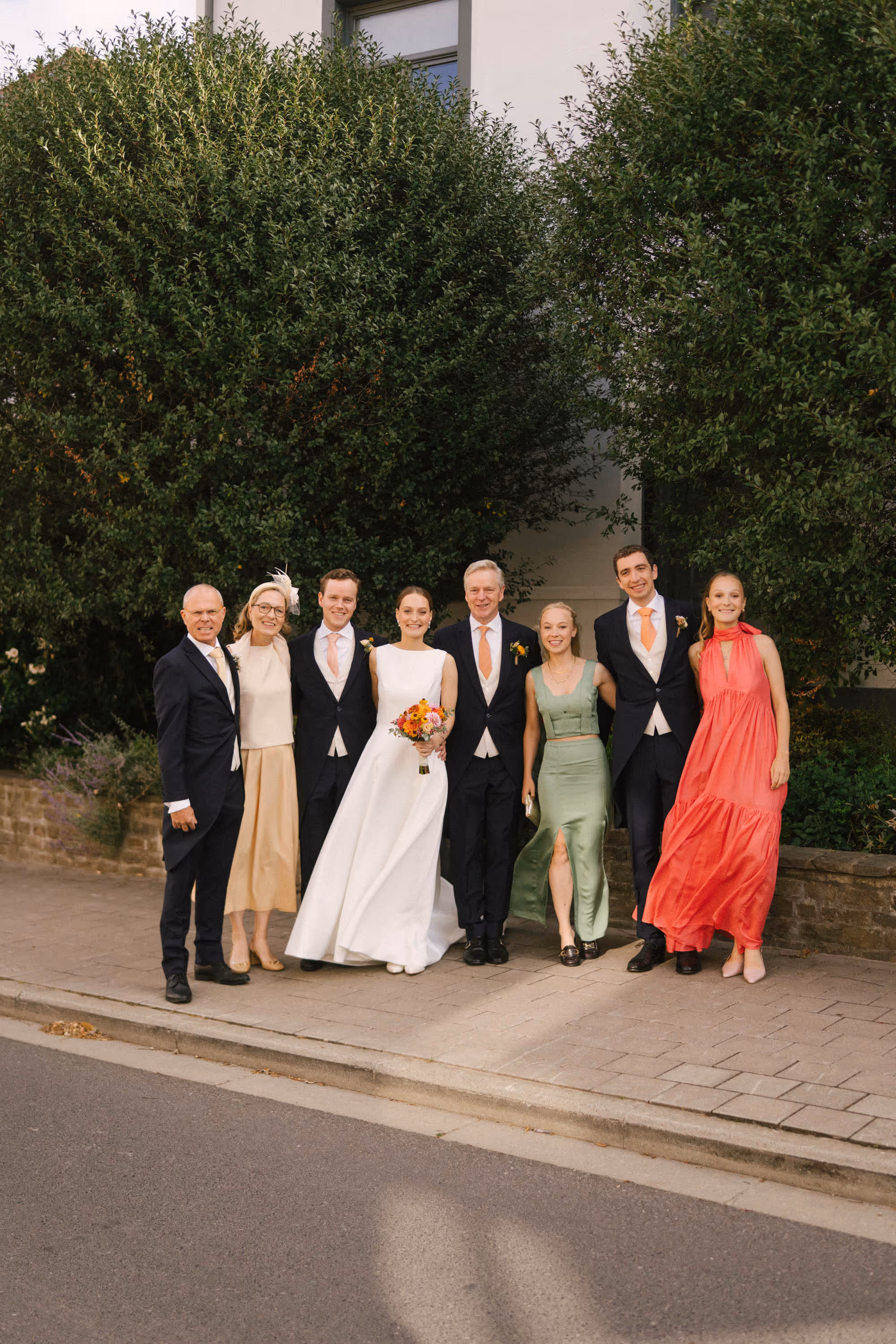 Seven people dressed formally posing on a sidewalk with greenery and a white building behind them, including a bride in a white dress holding a bouquet.