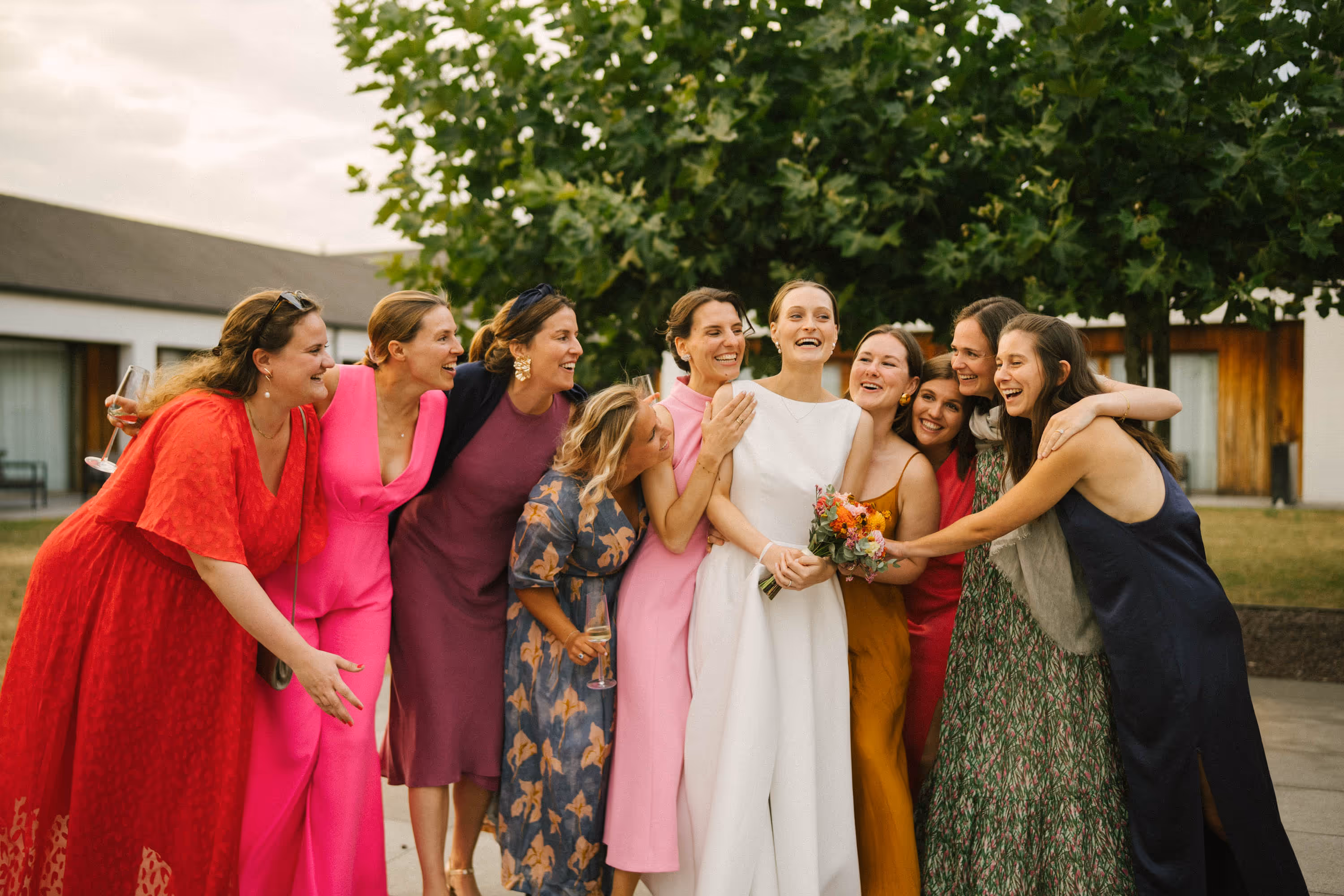 Group of nine women, including a bride in a white dress holding a bouquet, smiling and embracing outdoors.