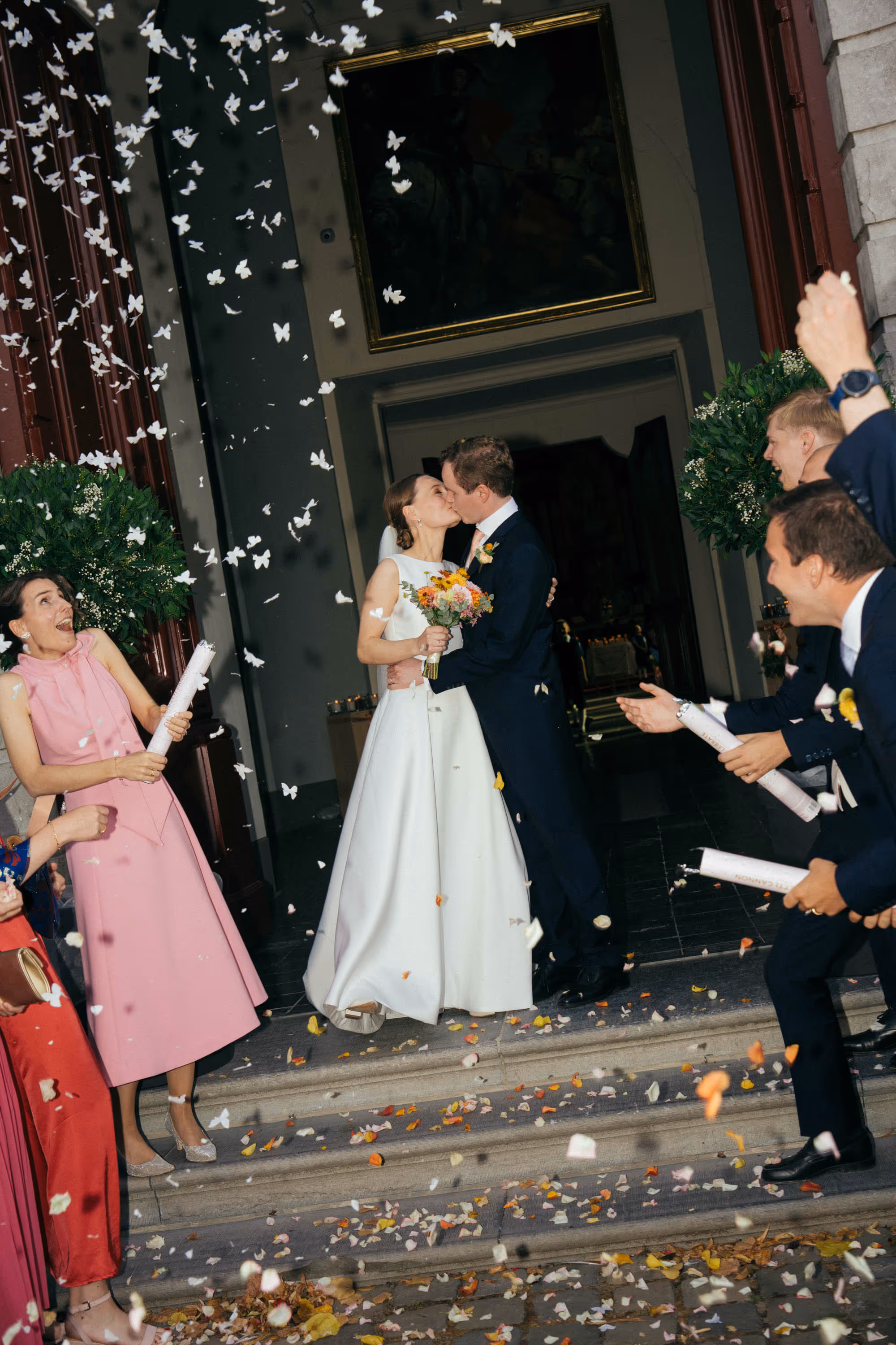 Bride and groom kissing on steps as guests celebrate by throwing rose petals.