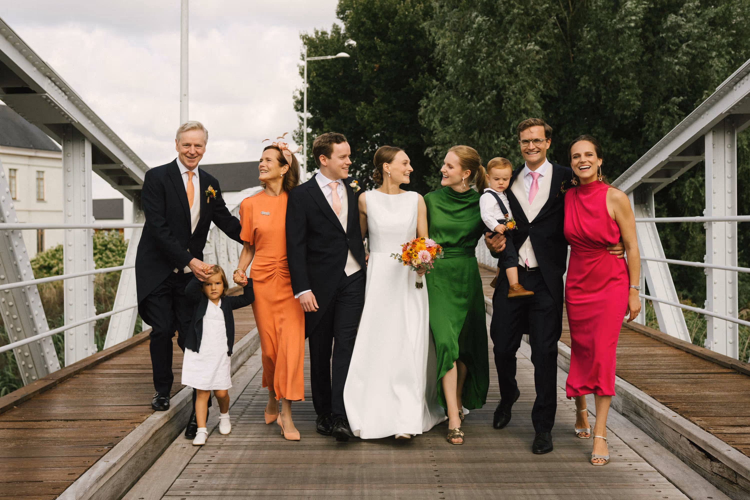 Group of well-dressed people including a bride in white holding a bouquet, standing on a wooden bridge with greenery in the background.