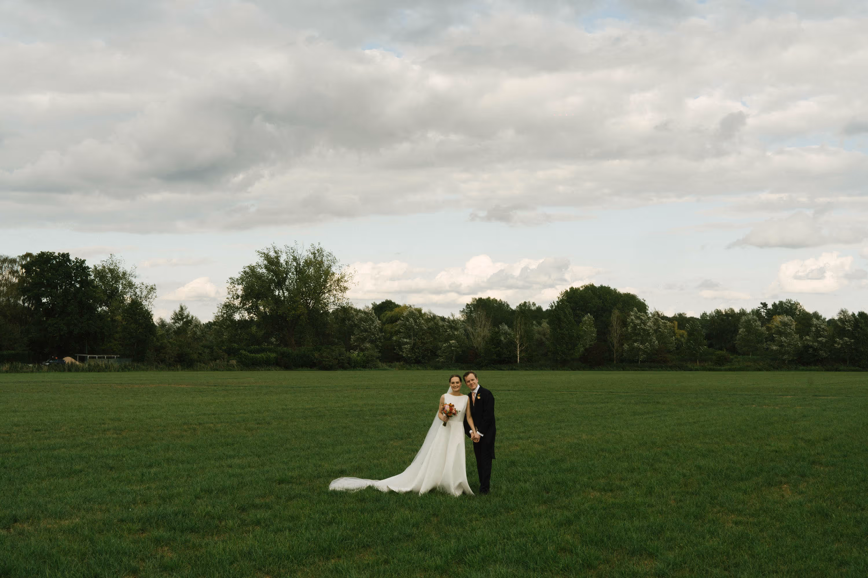 Bride in white gown and groom in black suit holding hands in a large green field with trees and cloudy sky in the background.