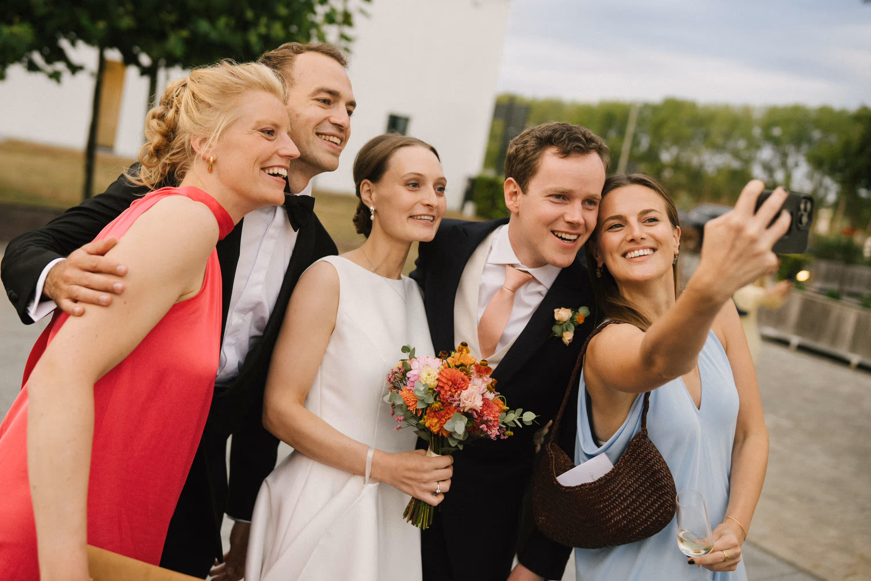 Group of five people smiling and posing for a selfie at a wedding, with the bride holding a colorful bouquet.