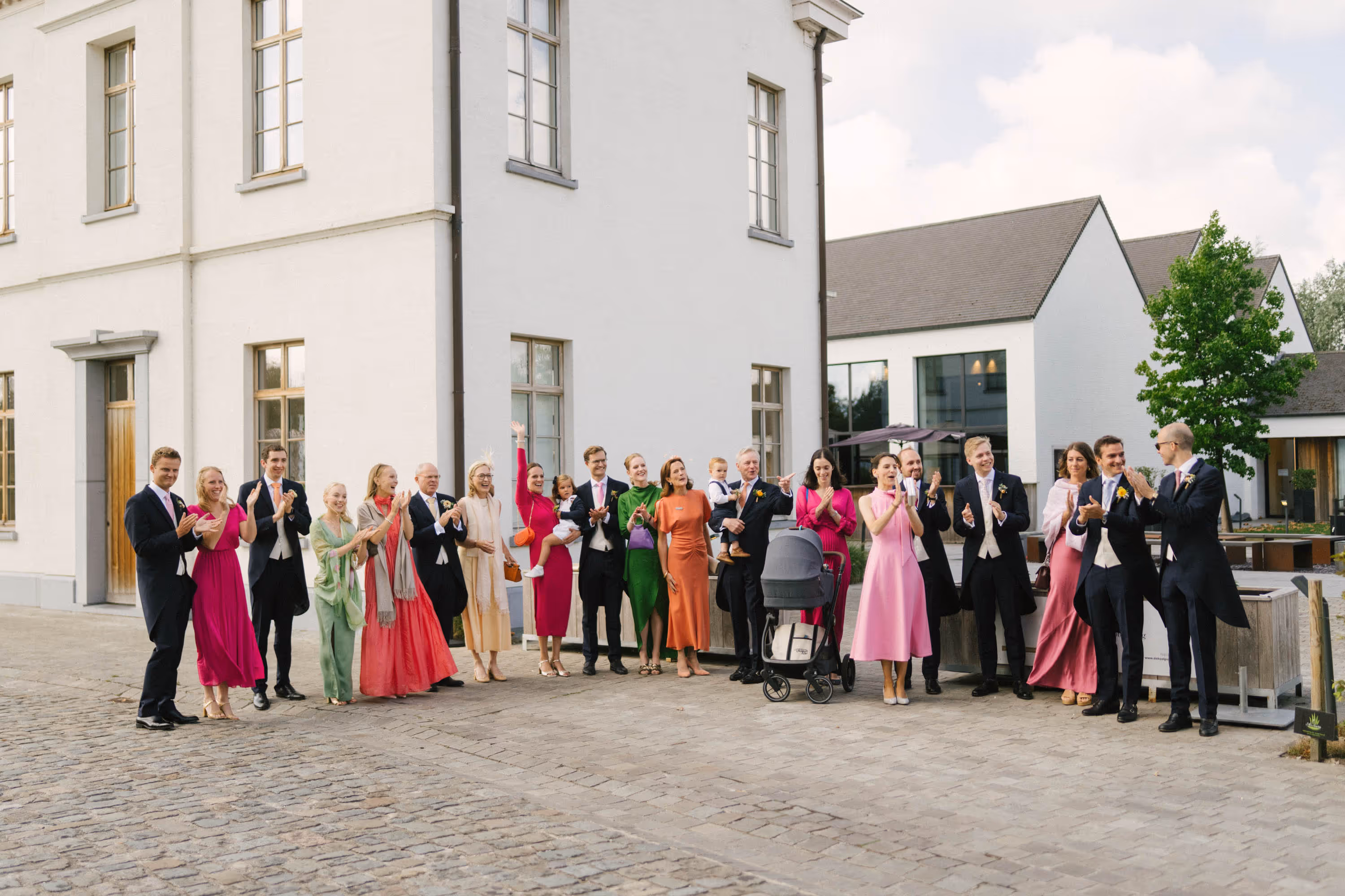 Group of well-dressed people clapping and smiling outside modern white buildings on a cobblestone street.