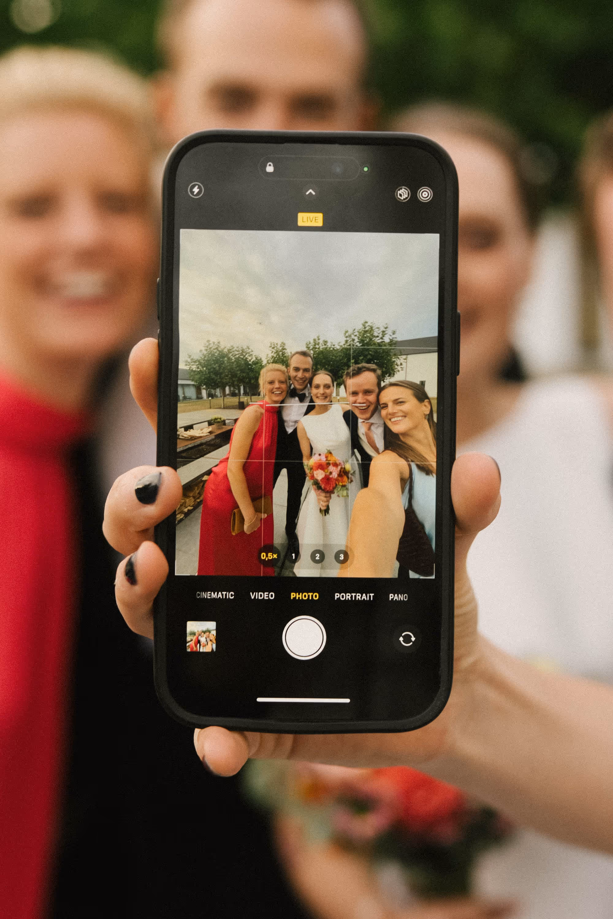 Group selfie of five people dressed formally, including a bride in a white dress holding a bouquet, shown on a smartphone screen held in front of the camera.