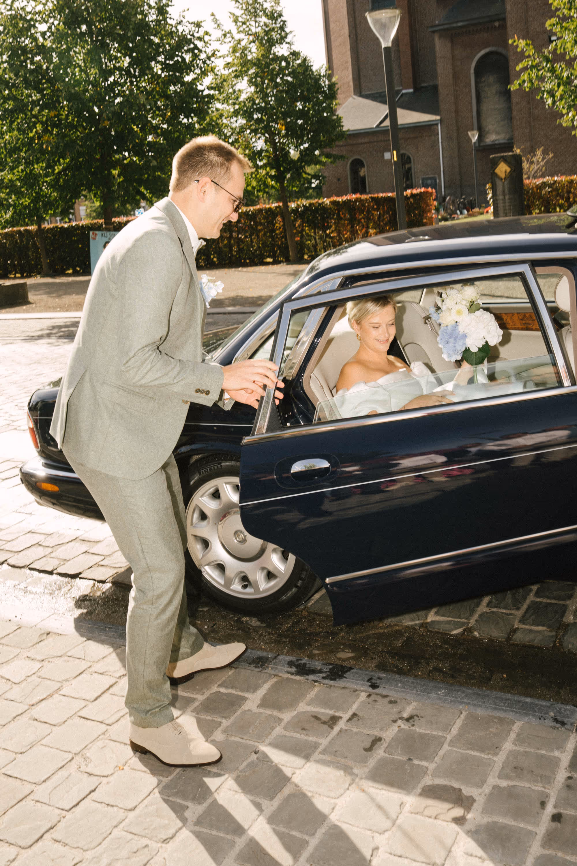 Man in light grey suit opening car door for woman in white dress holding bouquet, outdoors.